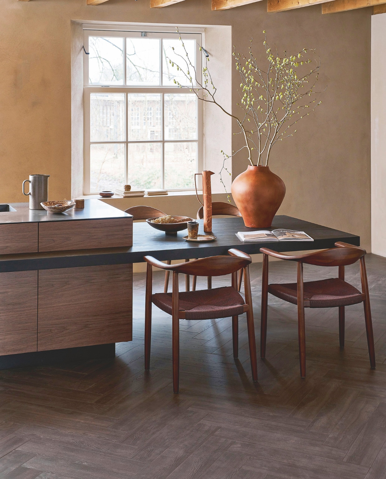 Laminaat visgraat in Collar Donkerbruin, lamel 65,5x13,3x0,8 cm, met groef, per m² € 37,99. M.m.v. shop.vtwonen.nl (eetkamerstoelen Embrace van vtwonen), mettesophie.com (hoge kan, aardewerken schaal), zarahome.com (bord), dofine.nl (wandafwerking), urbannatureculture.com (houten schaal, vaas). Overig privébezit. vtwonen 05-2025 | styling Cleo Scheulderman | fotografie Jeroen van der Spek