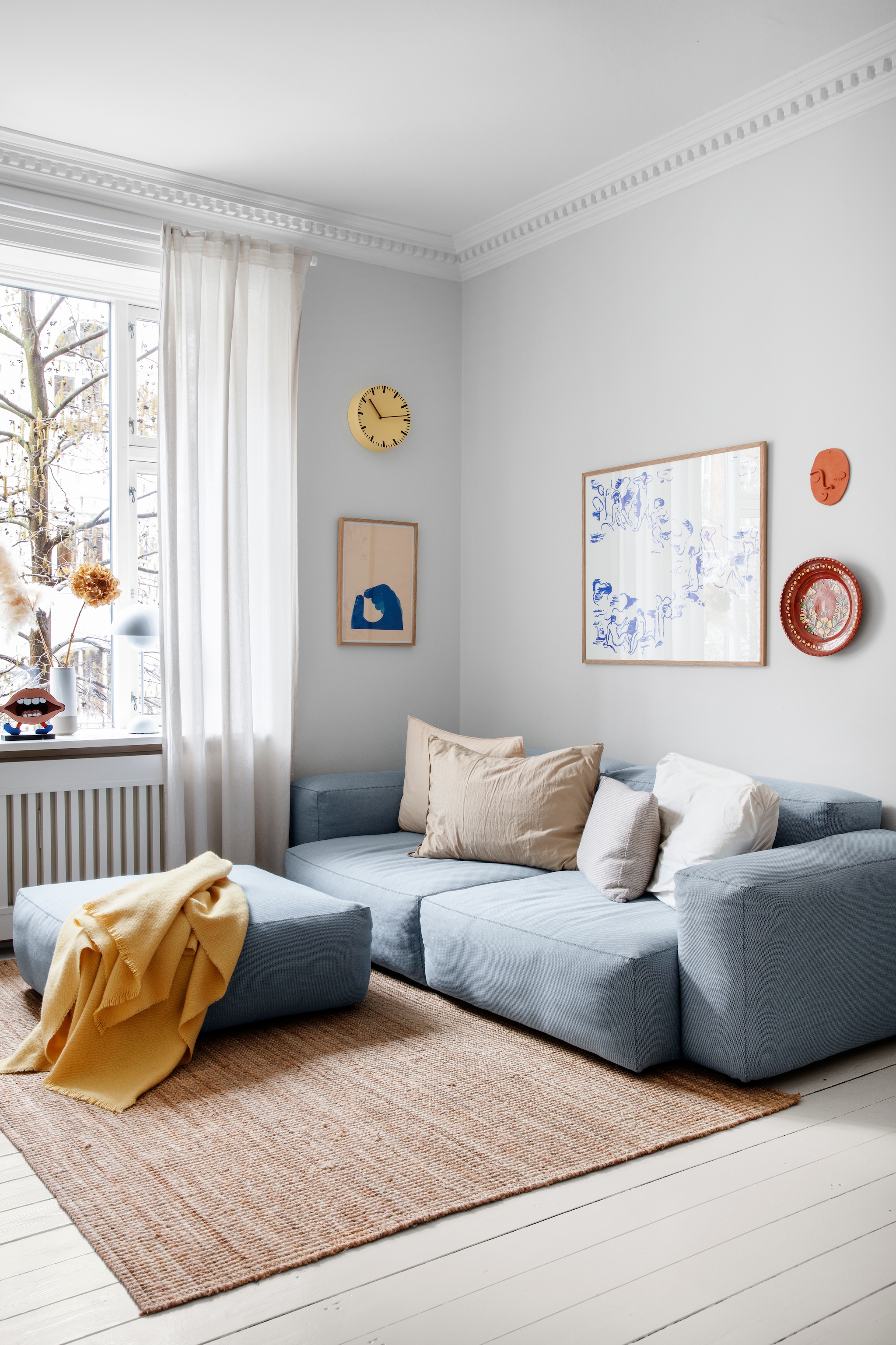 Bank, plaid en klok Hay. Kussens Aiayu. Lamp Flower pot van Verner Panton. Mr. Smiley van Troels Trier. Ingelijst behang ontwerp van Poul Henningsen. Keramieken object gezicht Emma Louise Gram Heegaard. Bord rommelmarkt. Vloerkleed en gordijnen Ikea. vtwonen 01-2026 | productie Living Inside | styling Rikke Graff Juel | fotografie Christina Kayser O.