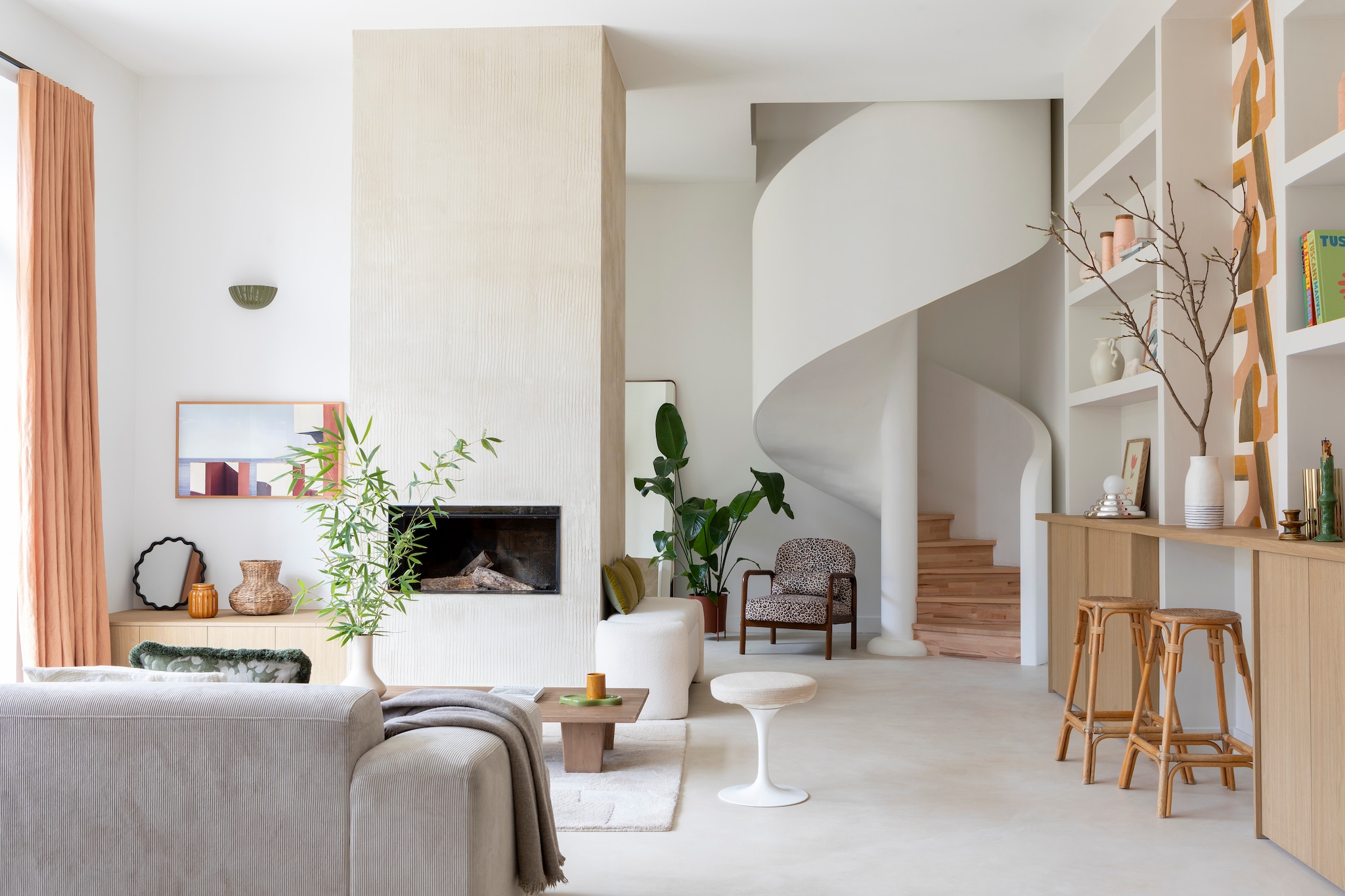 Staken trapstructuur, gipsen omkasting en beukenhouten treden Rouveure Márquez. Betonvloer en schouw Tlmt. Muurverf Porcelaine van Ressource Peintures. Inzethaard Flammes et Créations. Kasten Unilin. Terra vaas, witte vaas, spiegel, dienblad en vaas met tak H&m Home. Pot en kaars Les composantes. Tv The Frame van Samsung. Tapijt en bank Westwing. Salontafel Zara Home. Vintage Knoll-kruk, zijdespoelen en barkrukken vlooienmarkt Les Puces du Canal. Plaids Maison de vacances. Kussens HKliving. Fauteuil Sweeek. Linnen gordijnen Élitis. Behang Gino van Casamance. Chromen lamp van Axel Chay voor Monoprix. Witte vaas op plank Ferm Living. vtwonen 01-2026 | styling Marie Maud Levron | fotografie Pierrick Verny | productie AC Scoffoni