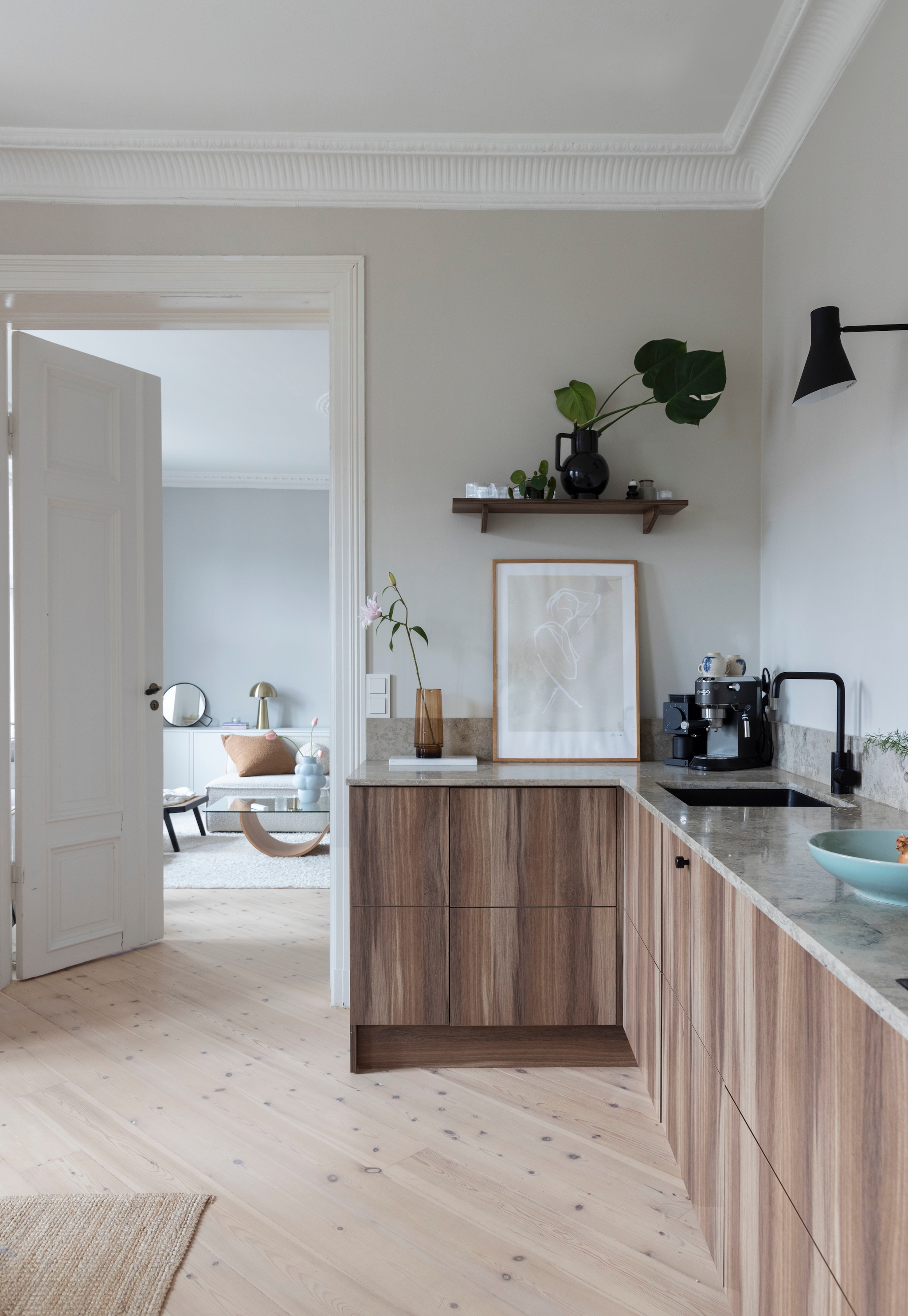 Keukenkasten in walnoot van een lokale keukenfirma. Grijs aanrechtblad in Jura-kalksteen. Spoelbak Ikea. Kraan tweedehands. Vergelijkbare wandlamp Vico van Olucia of AJ van Arne Jacobsen voor Louis Poulsen. Wandkleur Kalk van Jotun. vtwonen 01-2026 | styling Mari K. Olsen | fotografie Filippa Tredal | productie House of Pictures