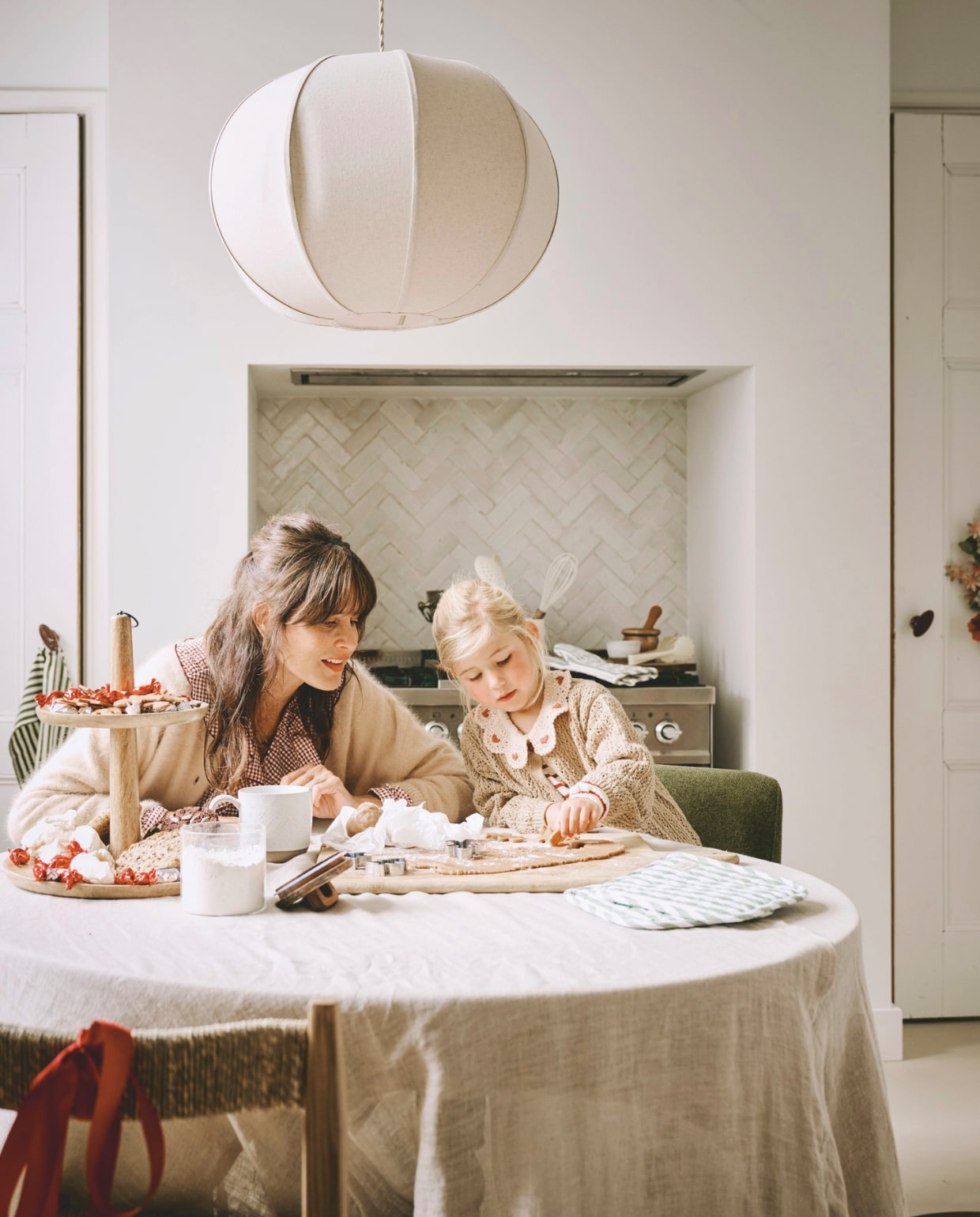 Zara Home tafelkleed, linnen € 69,99, stoel, essenhout en zeegras, 43x43x82 cm (bxdxh) € 189,-, hanglamp, katoen, wit, 48x24,5 cm (Øxh) € 79,99, voorraadpot met deksel, glas € 17,99, mok, keramiek, wit € 9,99, garde, kunststof € 9,99, pollepel, kunststof € 9,99, pot, keramiek, wit € 12,99, vijzel, hout € 19,99 (zarahome.com). H&m pannenlappen, groen-wit gestreept, set van 2 € 9,99, theedoek, groen-wit gestreept € 4,99, ovenwant, groen-wit gestreept € 10,99 (hm.com). The Green Home Company etagère, hout € 36,- (shop.vtwonen.nl). Polspotten eetkamerstoel Cosy, forest green, 57x63x83 cm (bxdxh) € 400,- (polspotten.nl). Bungalow bloemenkrans, metaal, verschillende kleuren € 80,- (bungalow.dk). Kleding Helena (moeder): Bellerose vest (orangebag.nl), Zara geruite top (zara.com), Massimo Dutti jeans (massimodutti.com). Kleding Saar (jongste dochter): Zara vest (zara.com), Lil’atelier jurk (lilatelier.com). vtwonen kerstspecial 2025 | styling Danielle Verheul | fotografie Sjoerd Eickmans