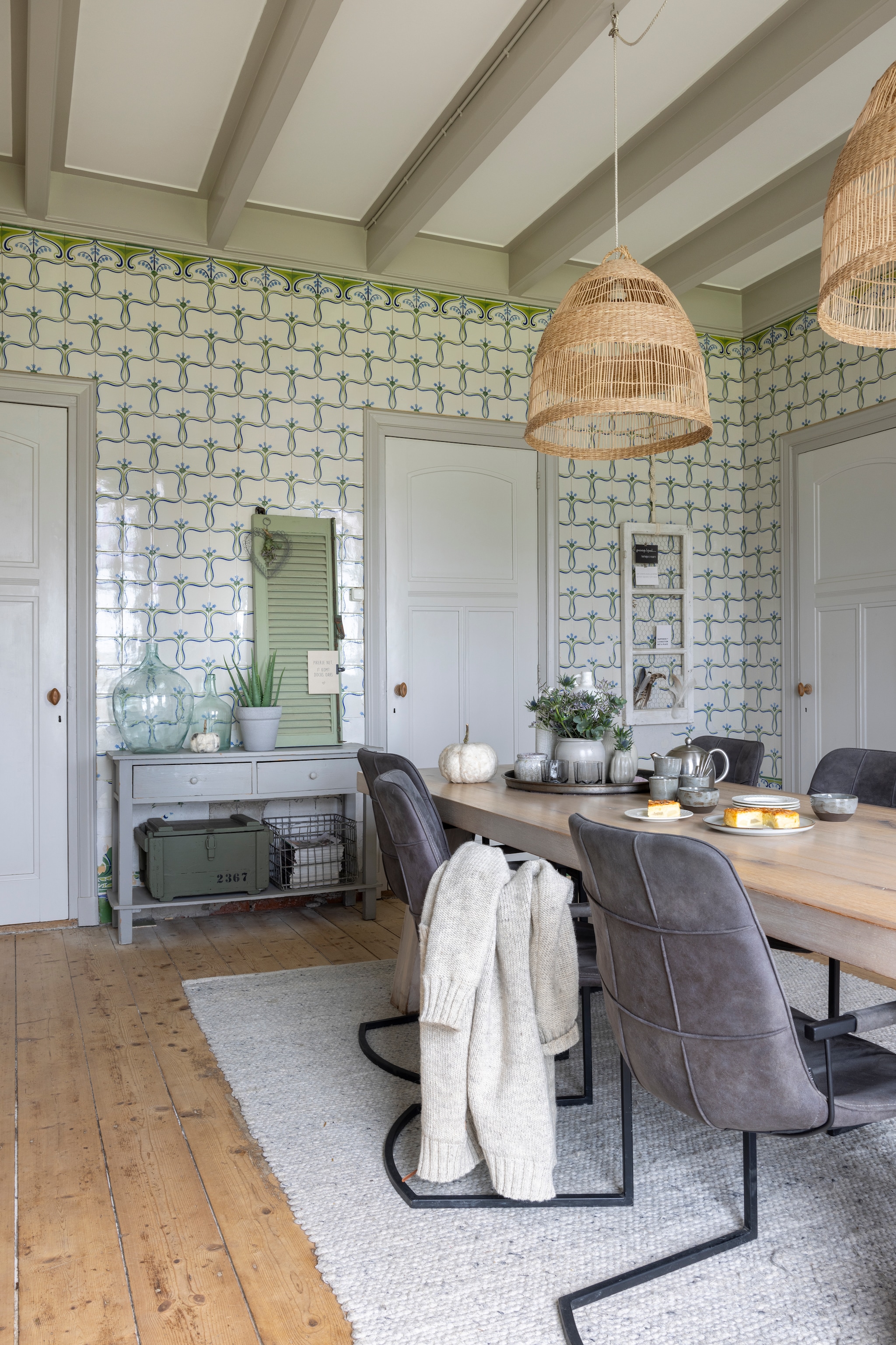 De eetkamer met de originele jugendstil tegelwand. Eettafel en sidetable Marktplaats. Vloerkleed Brinker Carpets. Rieten hanglampen Ikea. Groen luik gevonden op zolder. Servies Pottenbakkerij de Diggel. Wollen pompoen WenKR8. vtwonen Landelijk 02-2026 | Fotografie Anneke Gambon