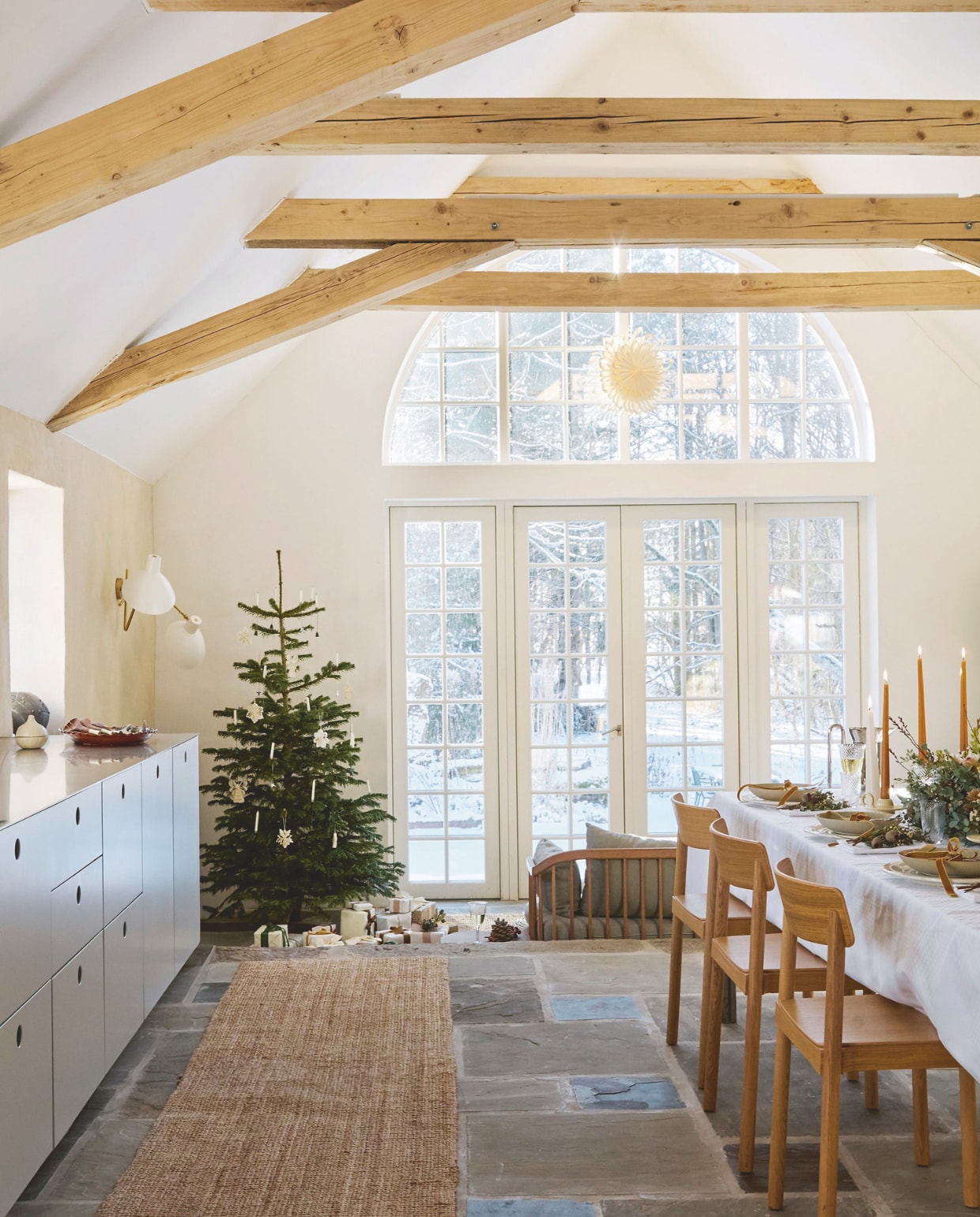 Cadeaupapier en lint Self Made. Wandlamp Astep. Bank Fly van Space Copenhagen. Ster voor het raam Sara Du Nord. Tafel ontworpen door Signe Hytte. Stoelen &Tradition. Boom versierd met origamisterren en echte kaarsen in houders. vtwonen 11-2025 | fotografie Glottipress