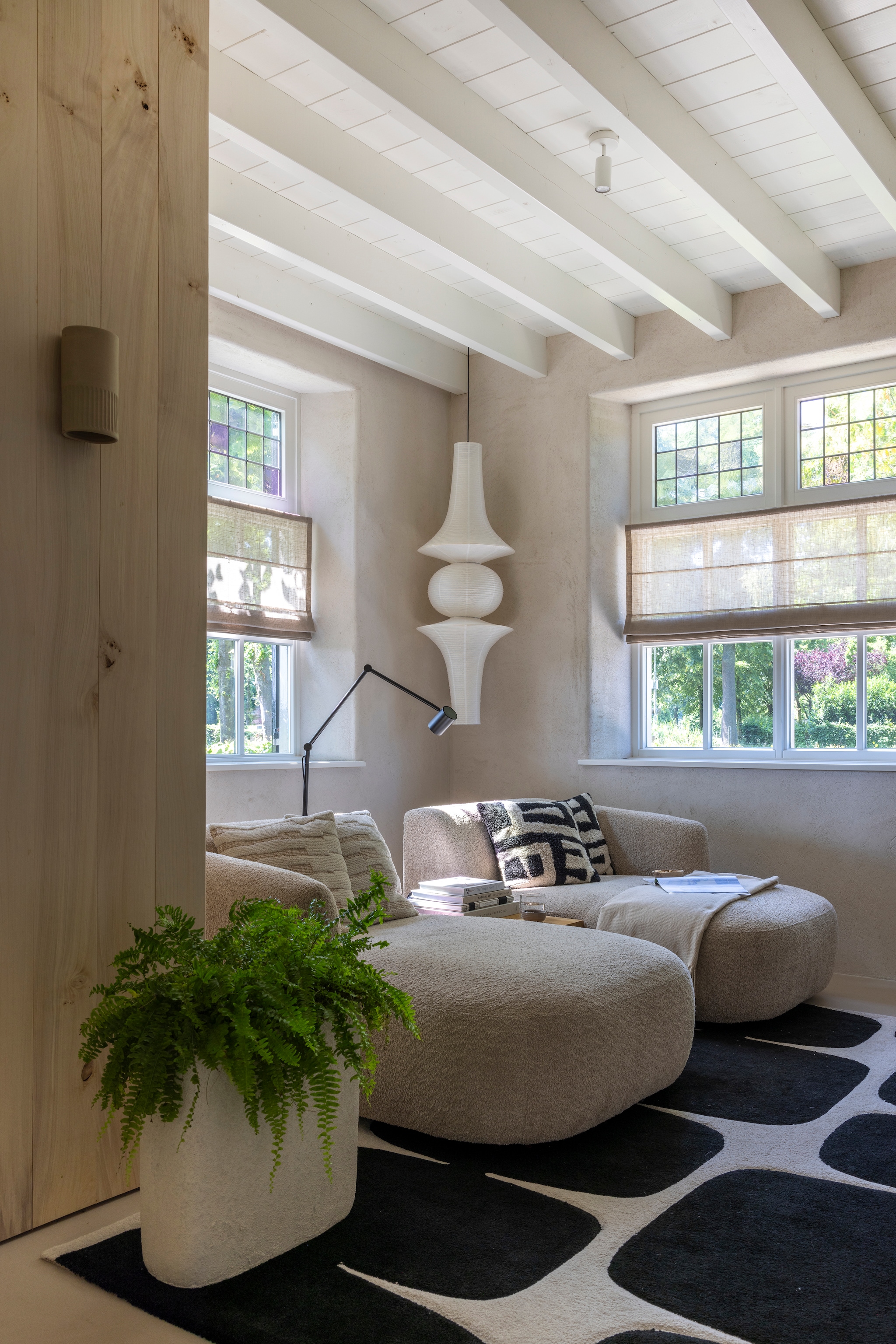 Banken Rozzano, kussens en vloerkleed Kwantum. Papieren hanglamp via Marktplaats. Wandlamp eigen ontwerp, uitgevoerd door Atelier Ikigai. Wand van populierenhout van Peppelhout. Vouwgordijnen Navajo 218442 van Raamstyling. Wanden leemstuc met leemfinish met toegevoegd stro kleur Kolumba Grau van Yosima. Het glas in lood is origineel en werd tussen dubbel glas gezet. vtwonen Landelijk 03-2026 | fotografie Anneke Gambon