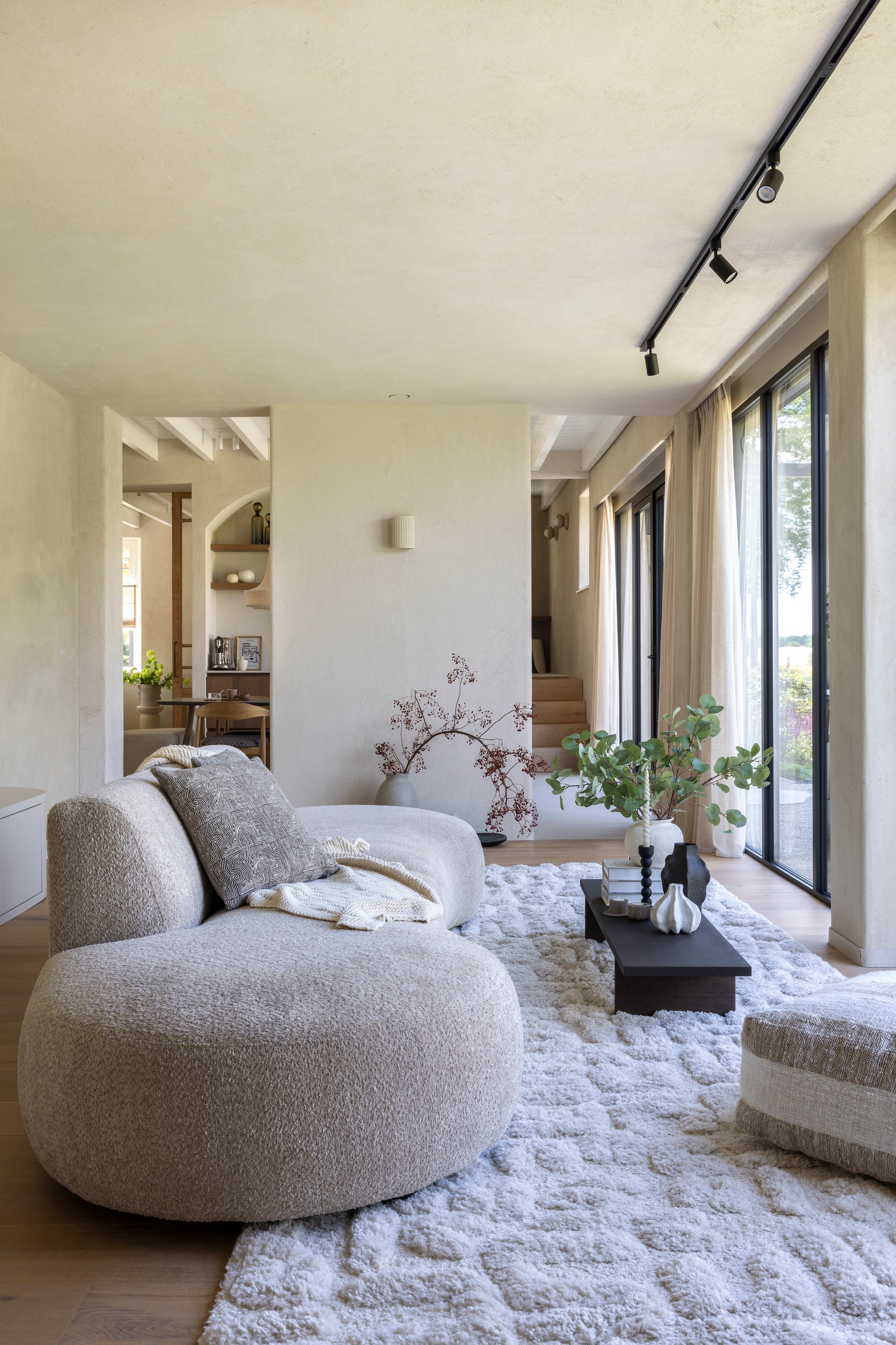 Bank Rozzano van Kwantum. Salontafel Kona Display van Ferm Living. Poef Coco homeliving. Vloerkleed Tibo van Benuta. Rail met spots Qub. Wandlamp eigen ontwerp, uitgevoerd door Atelier Ikigai. Gordijnen Navajo 218440 van Raamstyling. Vloerplanken in eiken 22 cm van Multiplank, afgewerkt met hardwasolie 2K Harea 029. Wanden witte basisleem van Tierrafino. Plafond leemfinish met toegevoegd stro kleur Kolumba Grau van Yosima.