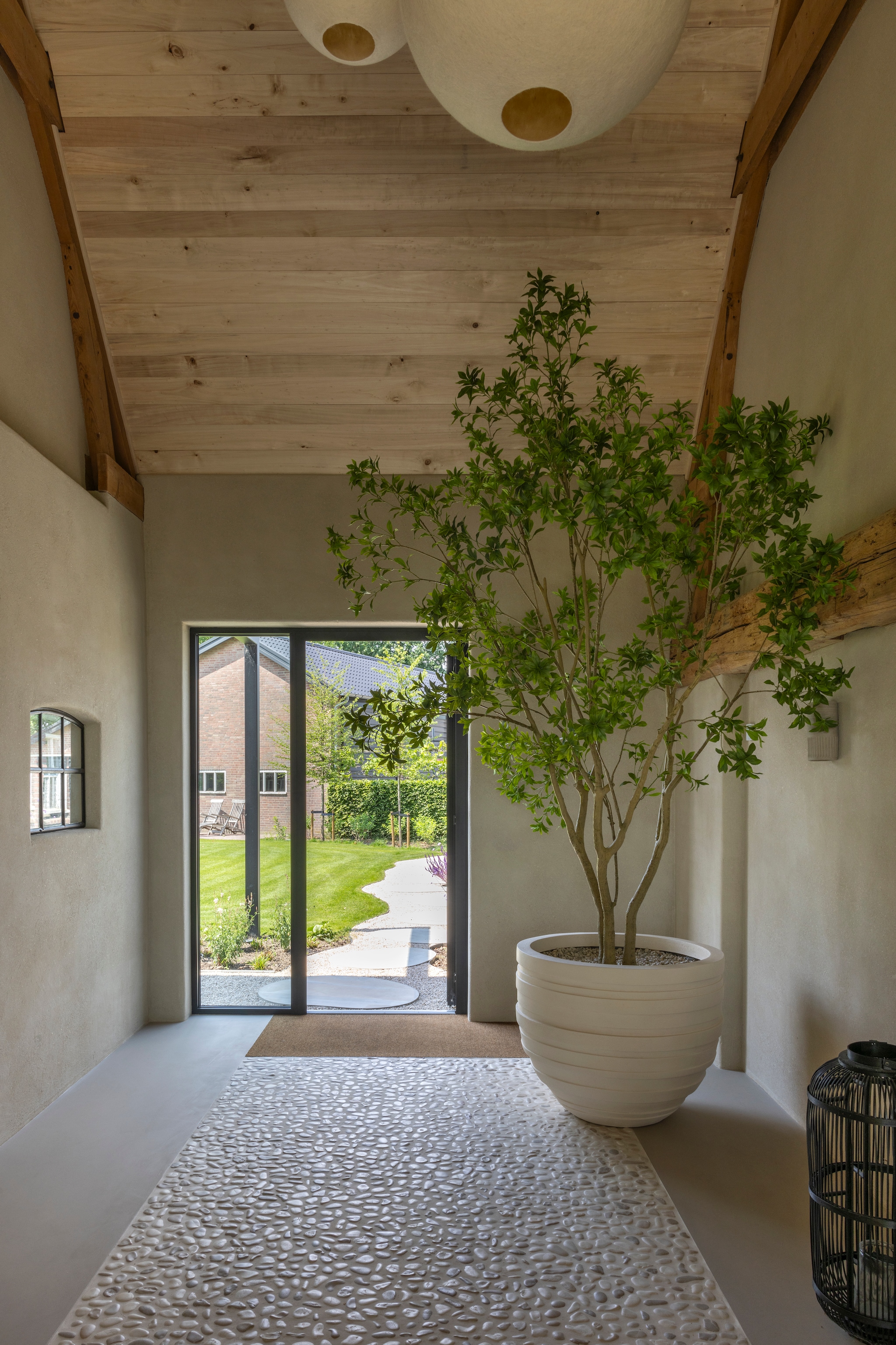 Pot met boom Emerald Eternal Green. Plafondpopulierenhout van Peppelhout. Lampen handgemaakt door Kalana Wool. Pebble-stenen via Pebble Shop. Vloer lavasteen van Van Pepijn. Wanden witte basisleem met toegevoegd stro van Tierrafino. vtwonen Landelijk 03-2026 | fotografie Anneke Gambon