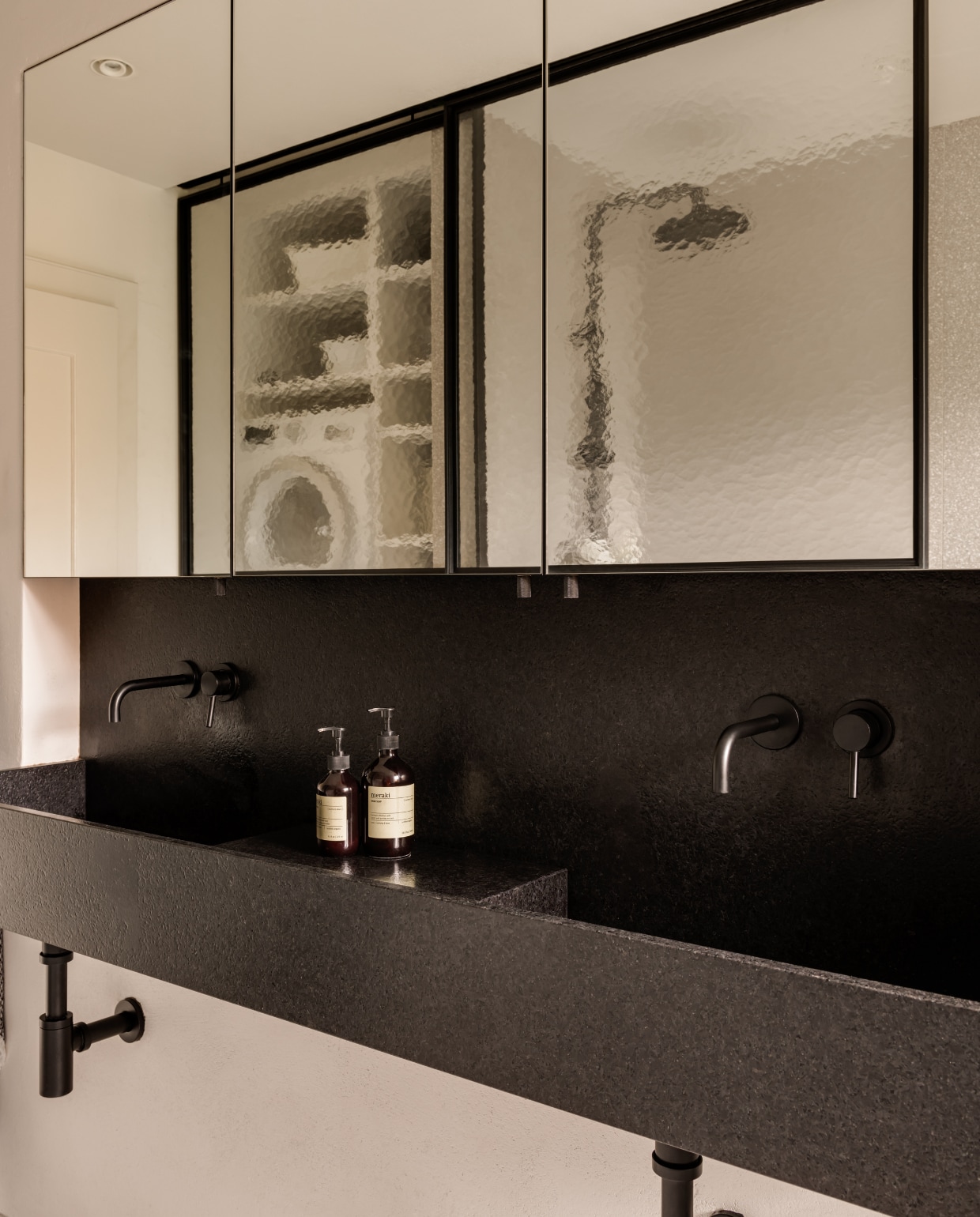 Grote wastafel van zwart graniet maatwerk, ontworpen door Piotr Kuchciński. Een wand van kathedraalglas schermt de douche en wasmachine af. stijlvol wonen 10-2025 | fotografie oni studio