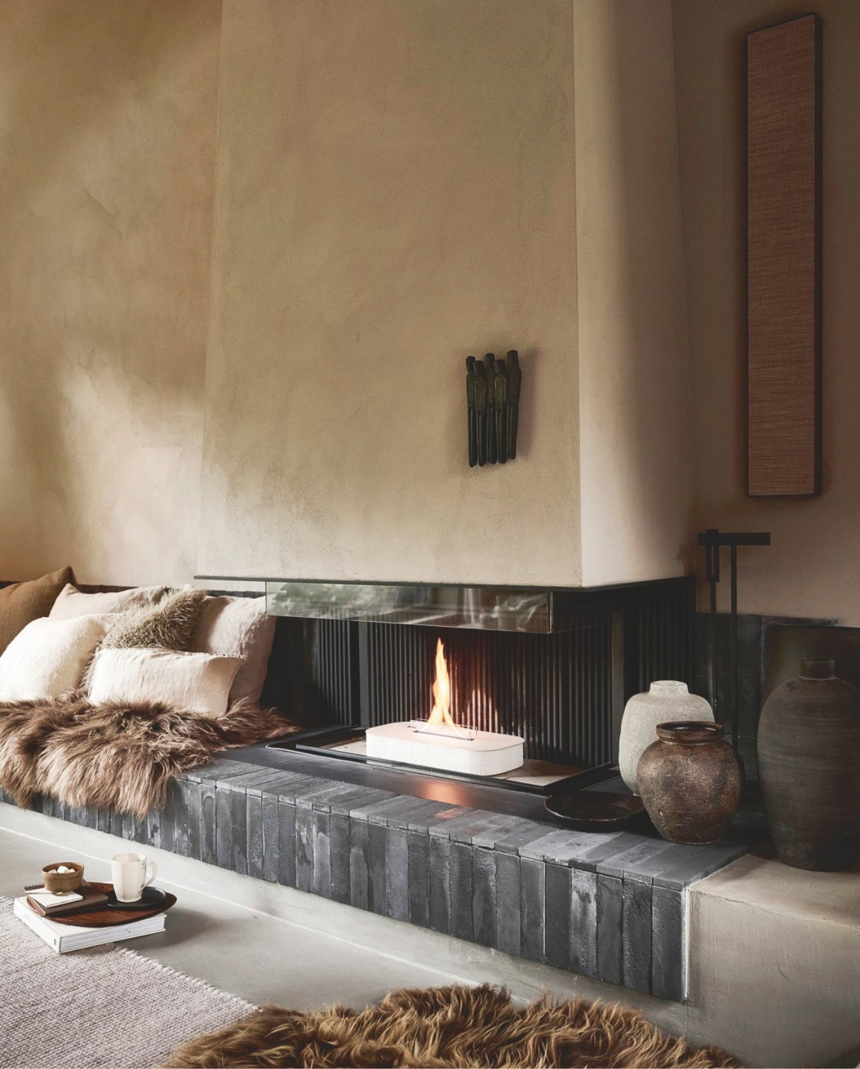 vtwonen tafelhaard Glow Aris 180, beton, beige, 47x22x12 cm (bxdxh) € 499,90.M.m.v. vachtenspecialist.nl (schapenvachten), fermliving.com (serveerplank), zarahome.com (schalen, vaas met structuur, bruin geweven kussenhoes, mok), hm.com (fluffy kussenhoes). Overig privébezit. vtwonen 12-2025 | styling sanne ketelaar | fotografie sjoerd eickmans