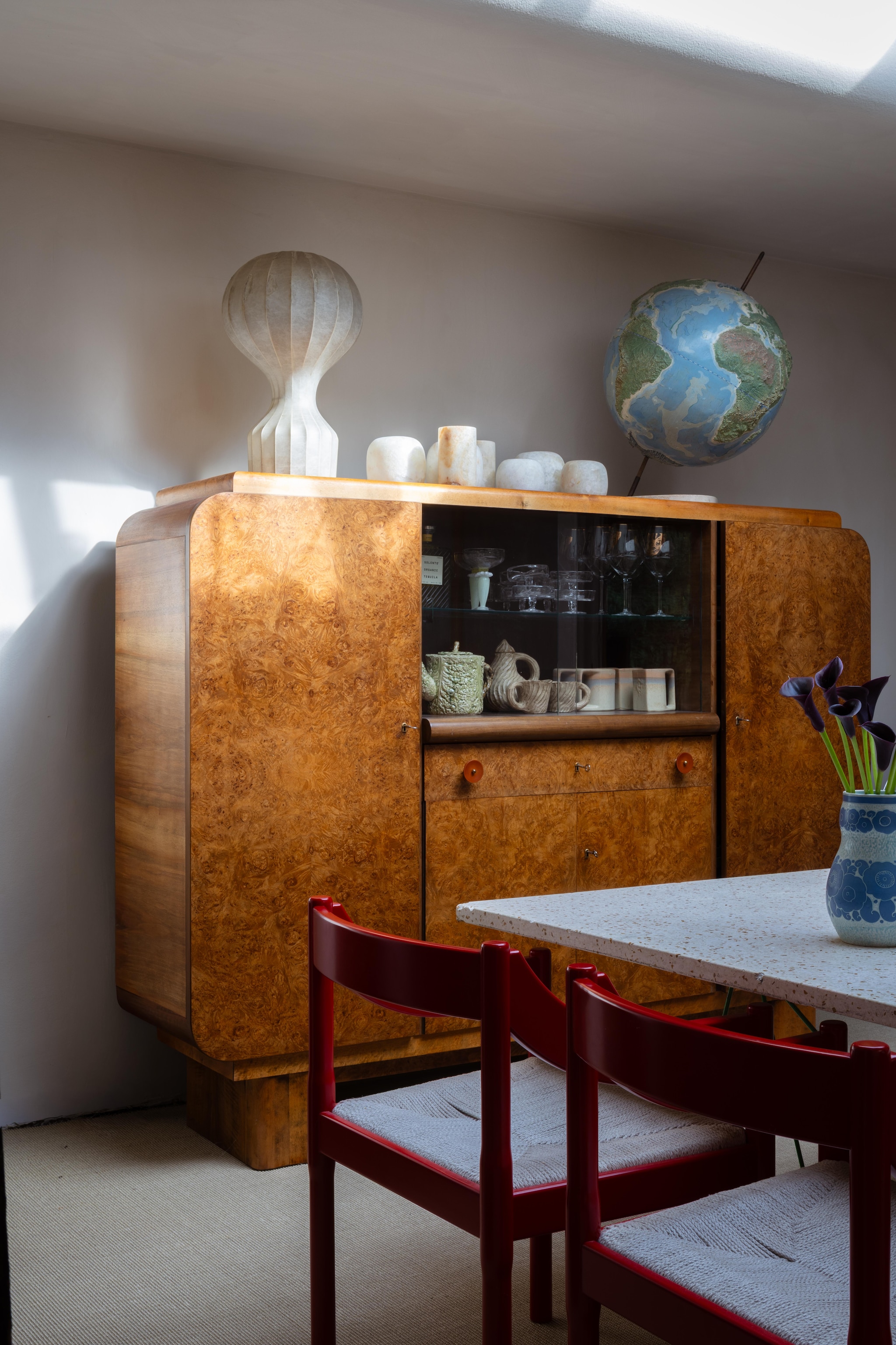 Antieke wandkast met objecten verzameld tijdens reizen en op markten. Tafellamp Cocoon van Flos vintage. Rode stoelen Magistretti. Vaas Rörstrand. vtwonen Design 03-2026 | fotografie Monica Spezia | productie Living Inside