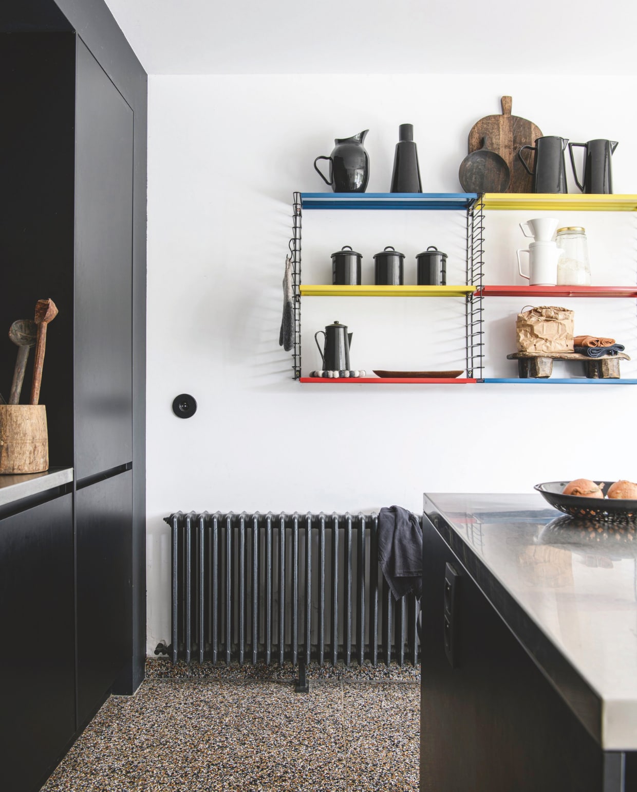 Keuken op maat laten maken door een timmerman. Radiator zat al in het huis, zwart geverfd. Terrazzo-tegelvloer uit Italië, via de aannemer. Wandrek, stopcontact, zwarte vaas, waterkannen, koffiepotten, drie zwarte potten en fruitschaal Zangra.