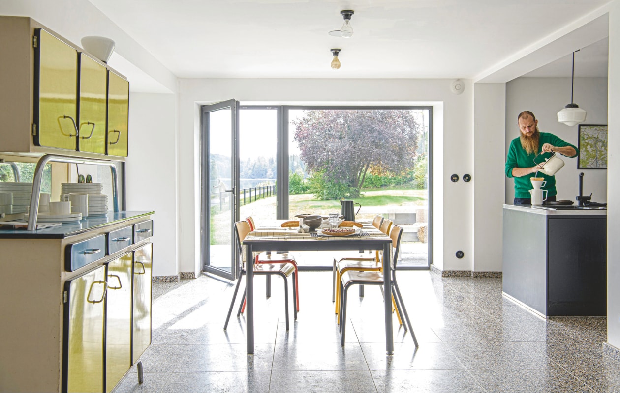 Keuken op maat laten maken door een timmerman. Terrazzo-tegelvloer uit Italië, via de aannemer. Geel-blauwe kast vintage. Eettafel gemaakt van oude bureaus. Stoelen, lichtschakelaars, stopcontact, lampen, koffiepot met filter en waterkan Zangra. vtwonen 12-2025 | styling Marit Saladini | fotografie Louis Lemaire