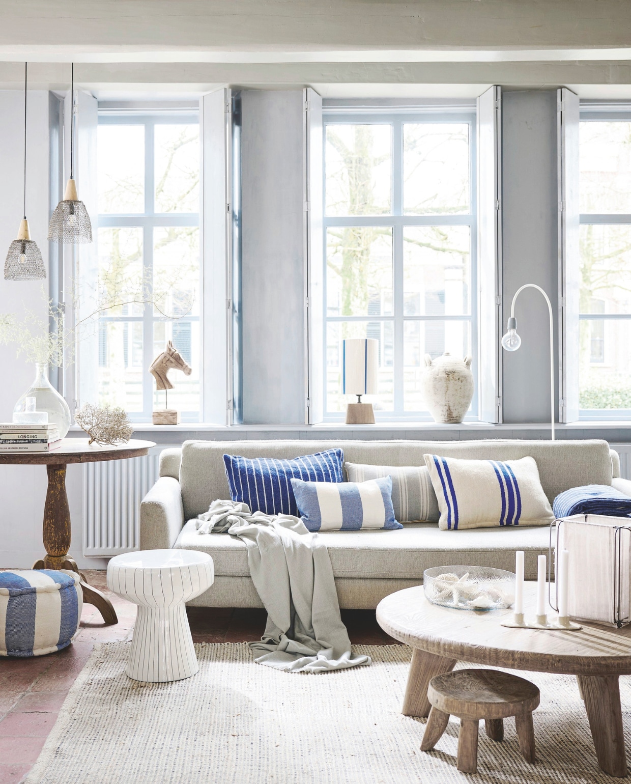 Oude houten salontafel Iker, 100x35 cm (Øxh) € 849,-, houten tafeltje uitIndia € 149,-, linnen windlicht € 59,95, metalen vloerlamp Concrete Bow van ETH, 157 cm (h) € 135,-, houten klassieke zuiltafel € 595,- (hetkabinet.nl). Katoenen gestreepte poef Navy, 35x20 cm (Øxh) € 99,- en wollen deken Pinstripe Midnight, vanaf 120x150 cm € 90,-  (thesouks.nl). Houten ornament Horse € 59,99, metalen bijzettafel Dakwa, 38x42 cm (Øxh) € 139,-, houten lampenvoet Kardan € 39,75 en jute draad lampenkap Medan, 20x30 cm (Øxh) € 51,99 van Light & Living, glazen kom Crinkle € 46,95 en glazen kandelaar Solvi B € 24,95 van Urban Nature Culture, vergelijkbare 2,5-zitsbank Rodeo van BePureHome, 190x86x85 cm (bxdxh) € 899,- (shop.vtwonen.nl). Kussens v.l.n.r.: wollen kussen Pinstripe Midnight € 70,-, gestreept katoenen kussen Navy € 50,- (thesouks.nl), kussen Coastal van HKliving, katoen, linnen en polyester € 49,95 (hetkabinet.nl) en wollen kussen Vintage streep 784 € 70,- (thesouks.nl). Glazen flesvaas € 29,95 (vandijkenko.nl). Gemêleerd katoenen jute vloerkleed, 200x300 cm € 219,-, keramieken kaarshouder € 17,99, lichtblauwe deken met franje, 140x190 cm € 39,99 (zarahome.com). Hanglampen, prijs op aanvraag (ushususthus.nl). Overig privébezit. vtwonen Landelijk 04-2025 | Styling Kim van Rossenberg | Fotografie Sjoerd Eickmans