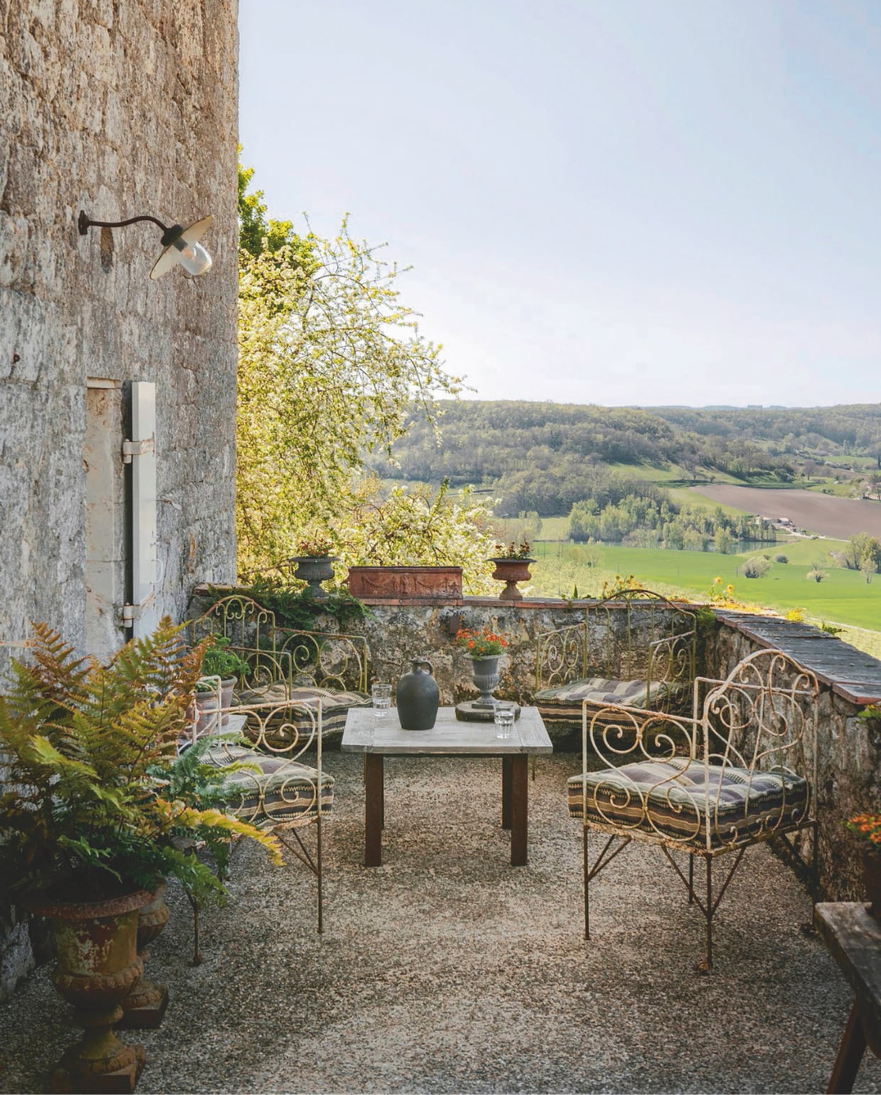 Het terras biedt een prachtig uitzicht op het omliggende landschap en het pittoreske plaatsje Penne-d’Agenais. Meubels en wandlamp vintage. Soortgelijke stoelen Beliani. vtwonen Landelijk 12-2025 | styling Amandine Berthon | fotografie Julien Fernandez