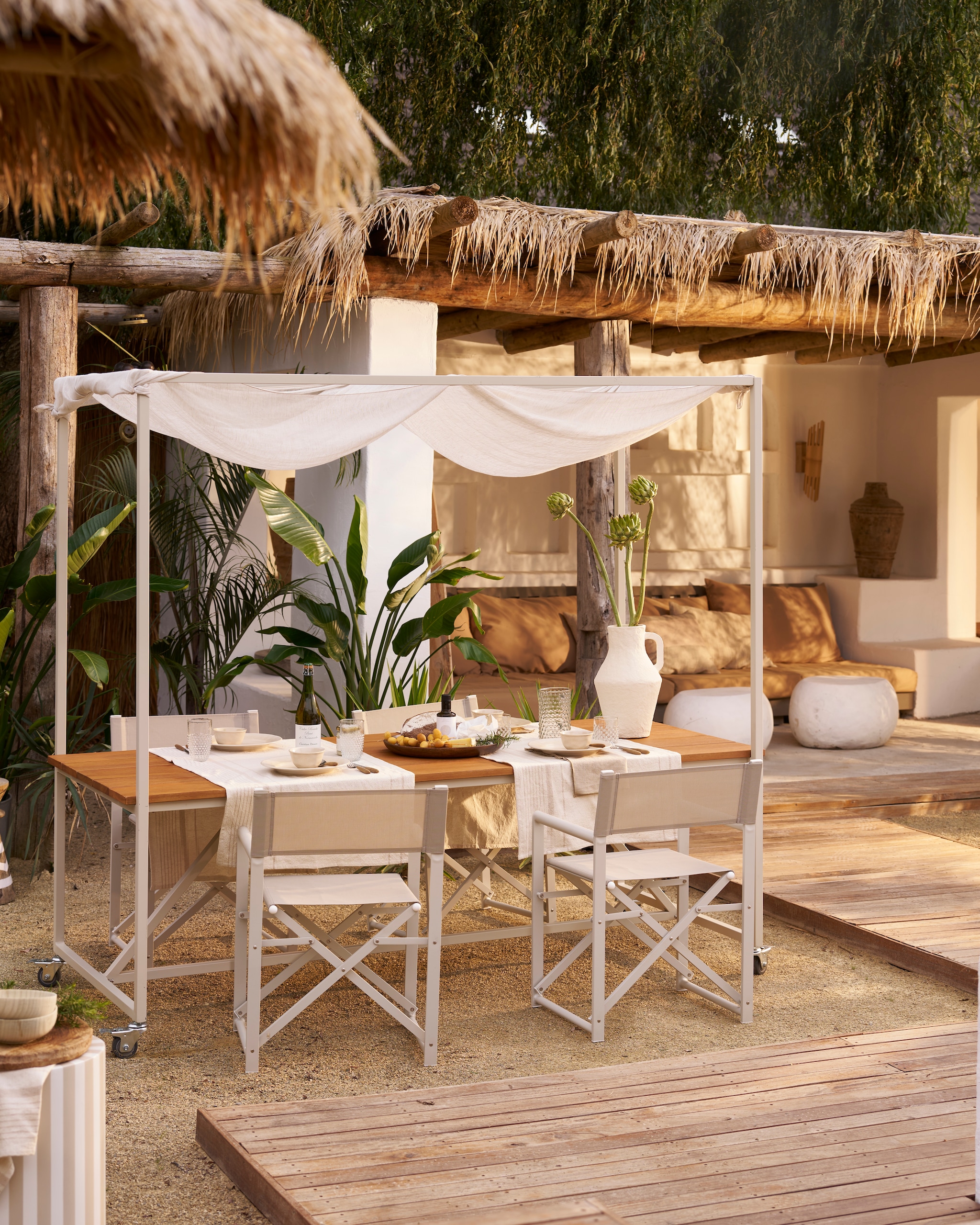 Eettafel Blue Sky op wielen, eucalyptushout en metaal, 220x200x97 cm (lxbxh) € 749,-, exclusief doek. Inklapbare regisseursstoel Director in Naturel, aluminium en stof, geschikt voor binnen en buiten, 56x49x81 cm (bxdxh) € 99,95. Fiberclay bijzettafels Pebble in betonlook wit, 65x49x31 cm (bxdxh) en 50x35 cm (Øxh), set van 2 € 378,-.M.m.v. vtwonen.nl/shop (glazen, karaf, tafellopers, houten schaal, gestreepte bijzettafel en kussens op buitenbank, allemaal van vtwonen), hm.com (servies), zarahome.com (vaas). Overig privébezit. vtwonen special 01-2026 | styling Cleo Scheulderman | fotografie Jeroen van der Spek