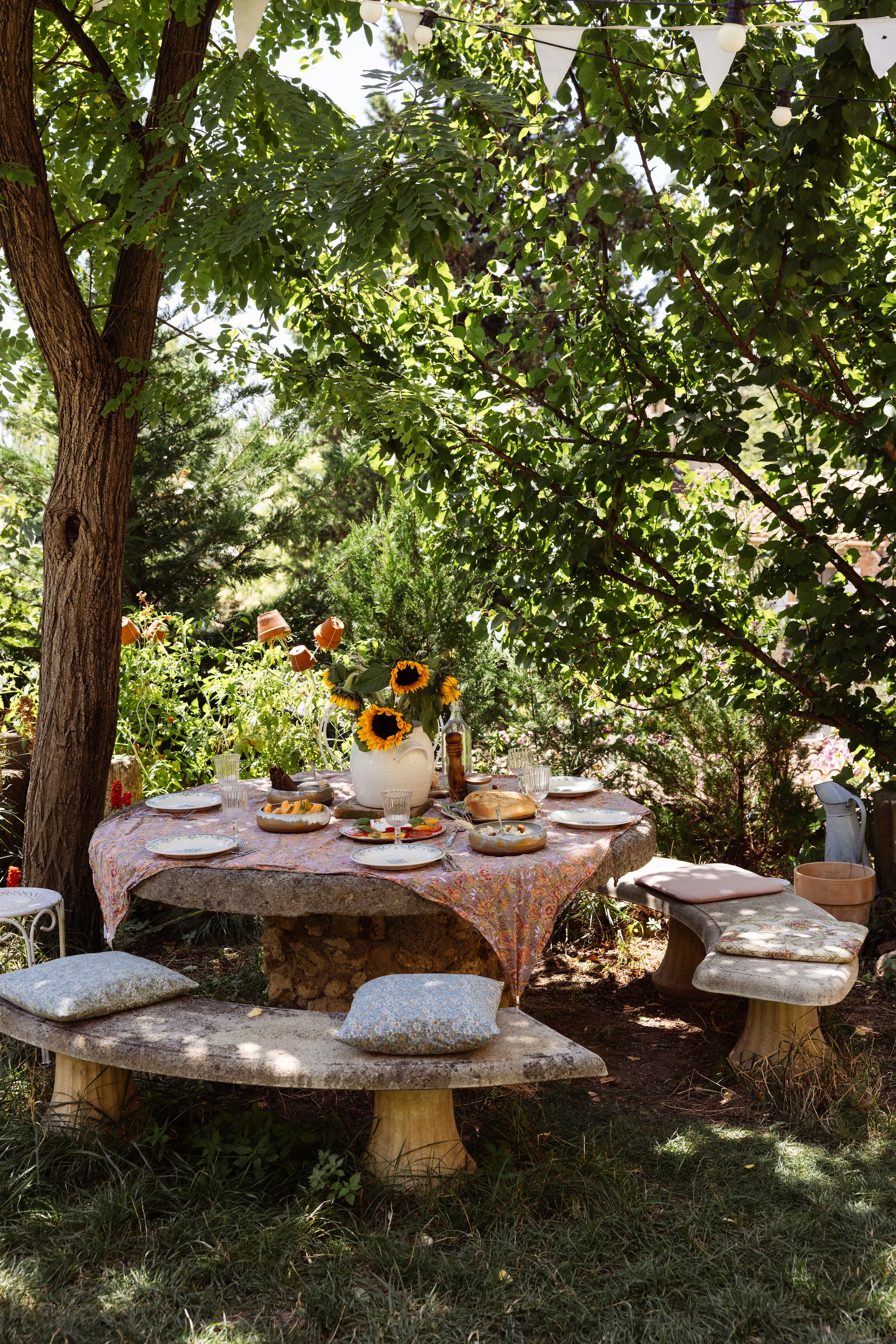 De oude, betonnen tafel met bankjes is een erfstuk. Tafelkleed Louise et Misha. Kussens zelfgemaakt. Oud servies en bestek Le French Finds. Grès pot brocante. vtwonen Landelijk 04-2026 | fotografie Dana van Leeuwen | styling Ilona Jongepier