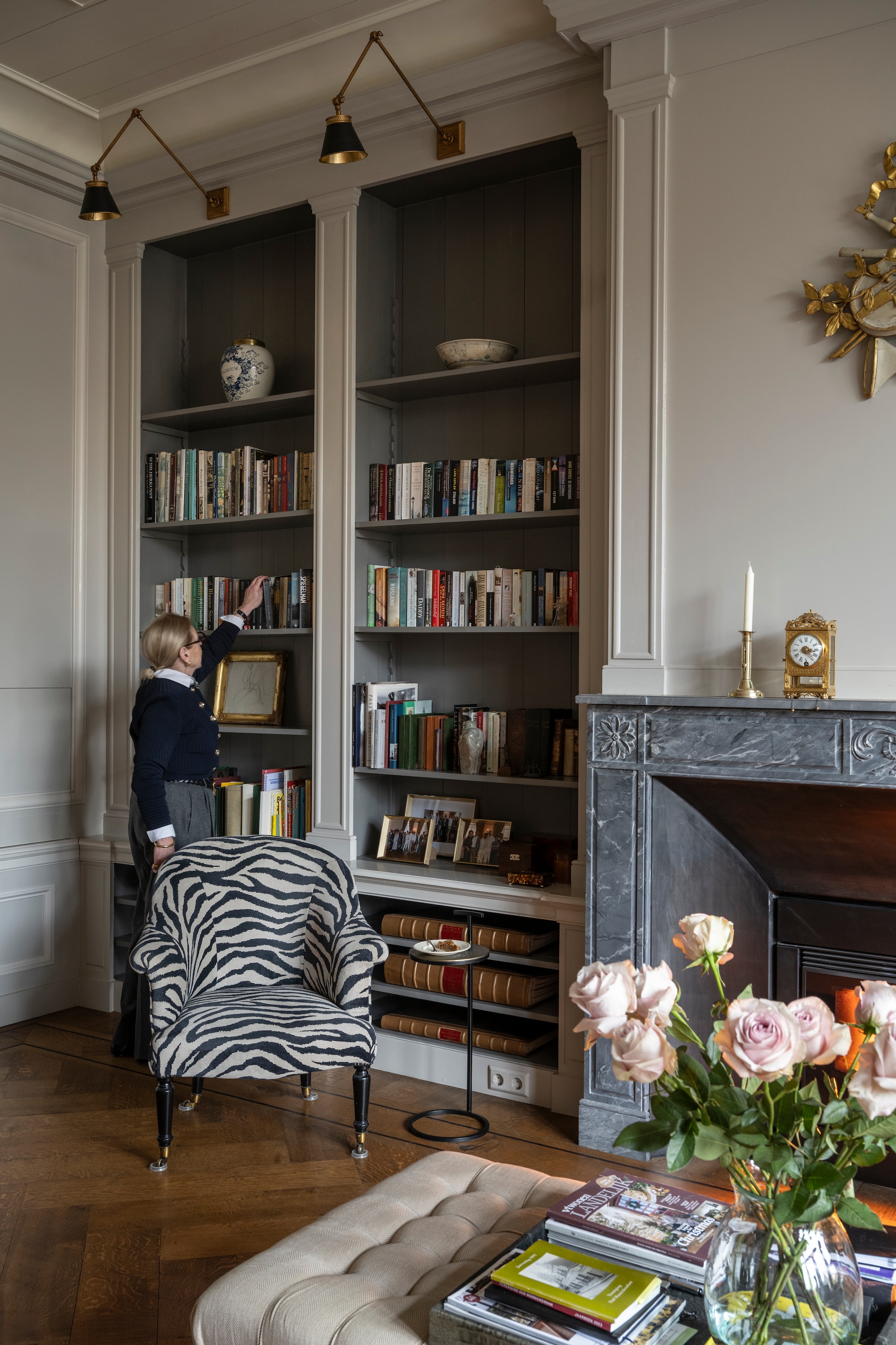 Muurkleur Purbeck Stone en kleur boekenkasten Mole’s Breath van Farrow & Ball. Wandlampen Empel Collections. Vazen, kandelaar, ornament aan de muur en schaal in de kast veilinghuis. Fauteuil met zebra-print bekleed door Ambiance. Bijzettafel Ambiance Home Decoration. Hocker Amelink. Klok De Klokkenmaker van Apeldoorn. Vloer Leguit en Roos. vtwonen Landelijk 04-2026 | styling Marianne Luning | fotografie Stan Koolen