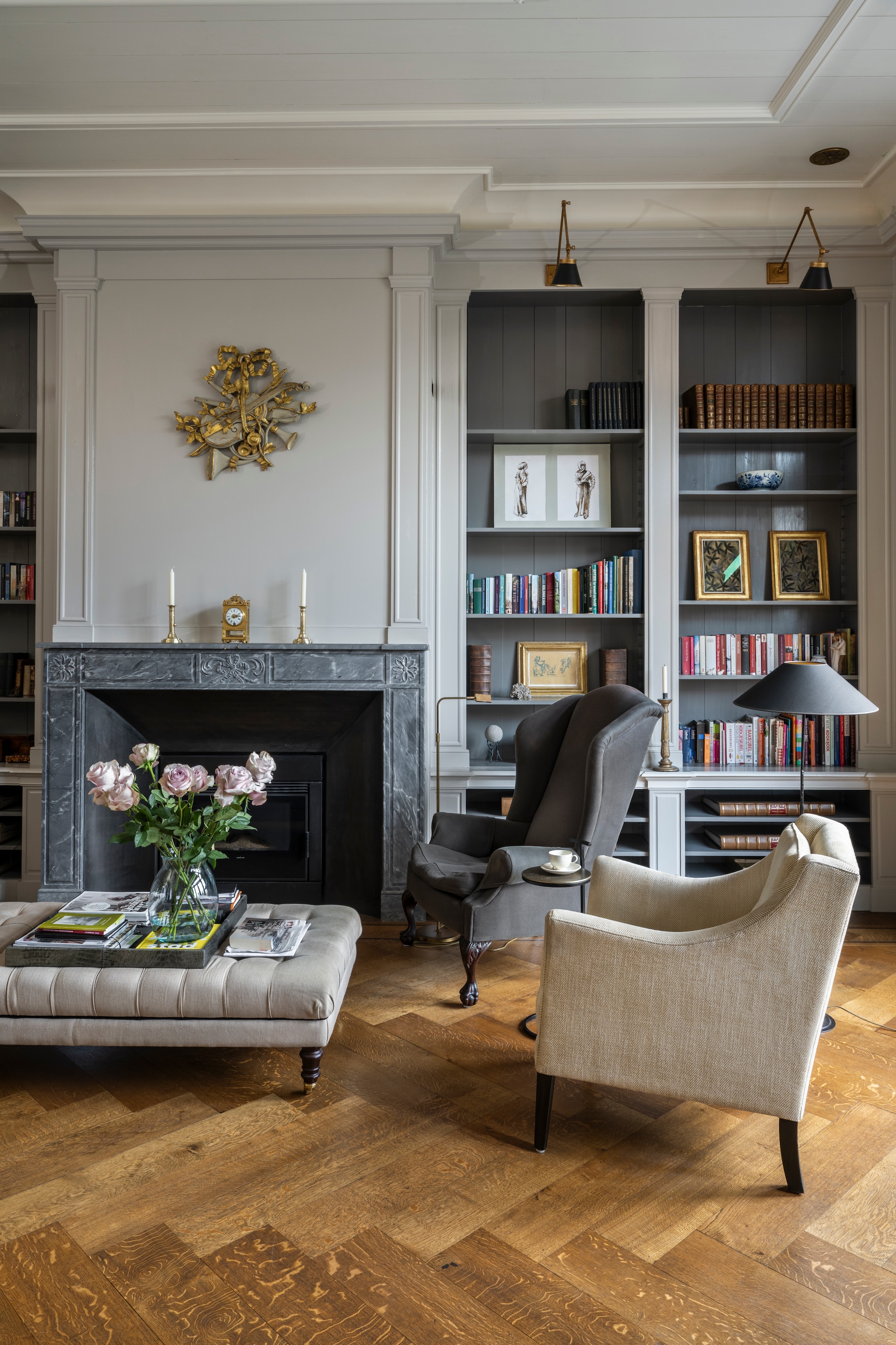 Beige fauteuil Dols & Co Interiors. Grijze fauteuil en stalamp Ambiance Home Decoration. Hocker Amelink. Muurkleur Purbeck Stone en kleur boekenkasten Mole’s Breath van Farrow & Ball. Wandlampen Empel Collections. Kunst in de kast Marjolein Juray. Klok De Klokkenmaker van Apeldoorn. Ornament aan de muur en kandelaars veilinghuis. Vloer Leguit en Roos. vtwonen Landelijk 04-2026 | styling Marianne Luning | fotografie Stan Koolen