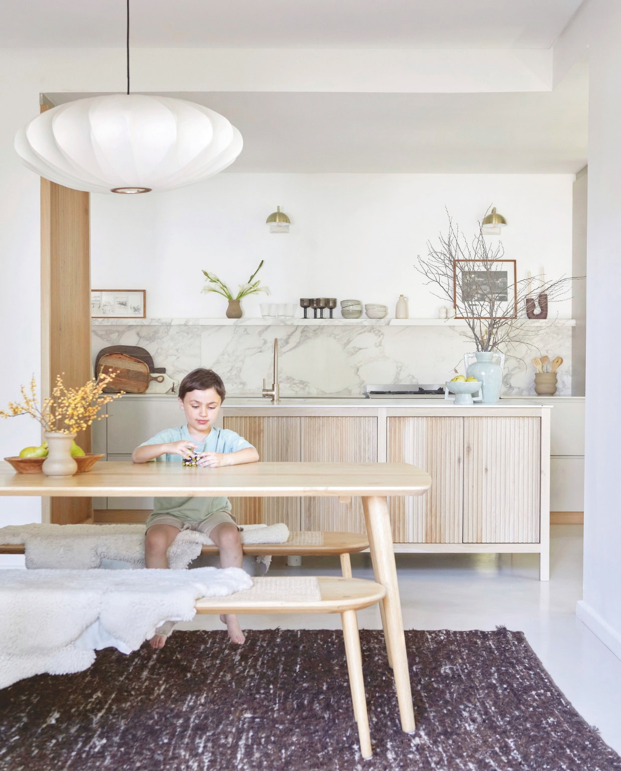 Keuken maatwerk, kookeiland bekleed met eikenhout. Composietstenen aanrechtbladen en spatbord met plank Infinity Surfaces. Muurlampen Coricraft. Combi gas-elektrisch fornuis Smeg. Kraan Meir Sa. Fruitschaal en vaas @home. Tafel en banken Houtlander. Hanglamp Spazio Lighting. Wollen vloerkleed Weylandts. Kunstwerk links Paul Senyol. Aardewerken pot Bernstein Pottery. Glazen en bakjes H&m Home. Kunstwerk rechts Sipho Mpongo. Kandelaar Pezula Interiors. vtwonen 02-2026 | styling Sven Alberding | fotografie Greg Cox