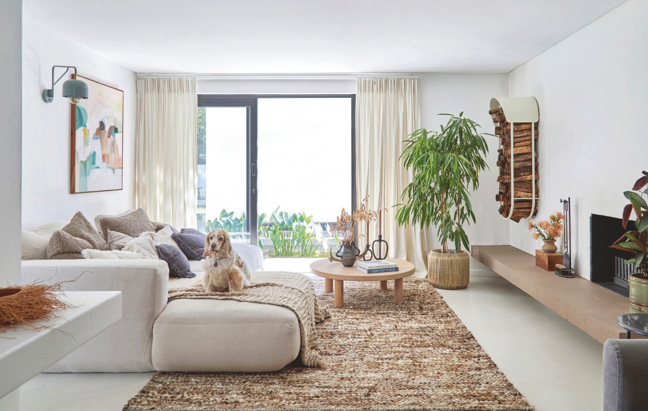 Bank La Grange Interiors. Muurlamp Acrobat van Acre Studio. Kunstwerk Laurinda Belcher. Vloerkleed, kussens en plaids Weylandts. Koffietafel Autumn and Oak. Kandelaars H&m Home. Vaas gemaakt door Janine voor Foraged. vtwonen 02-2026 | styling Sven Alberding | fotografie Greg Cox