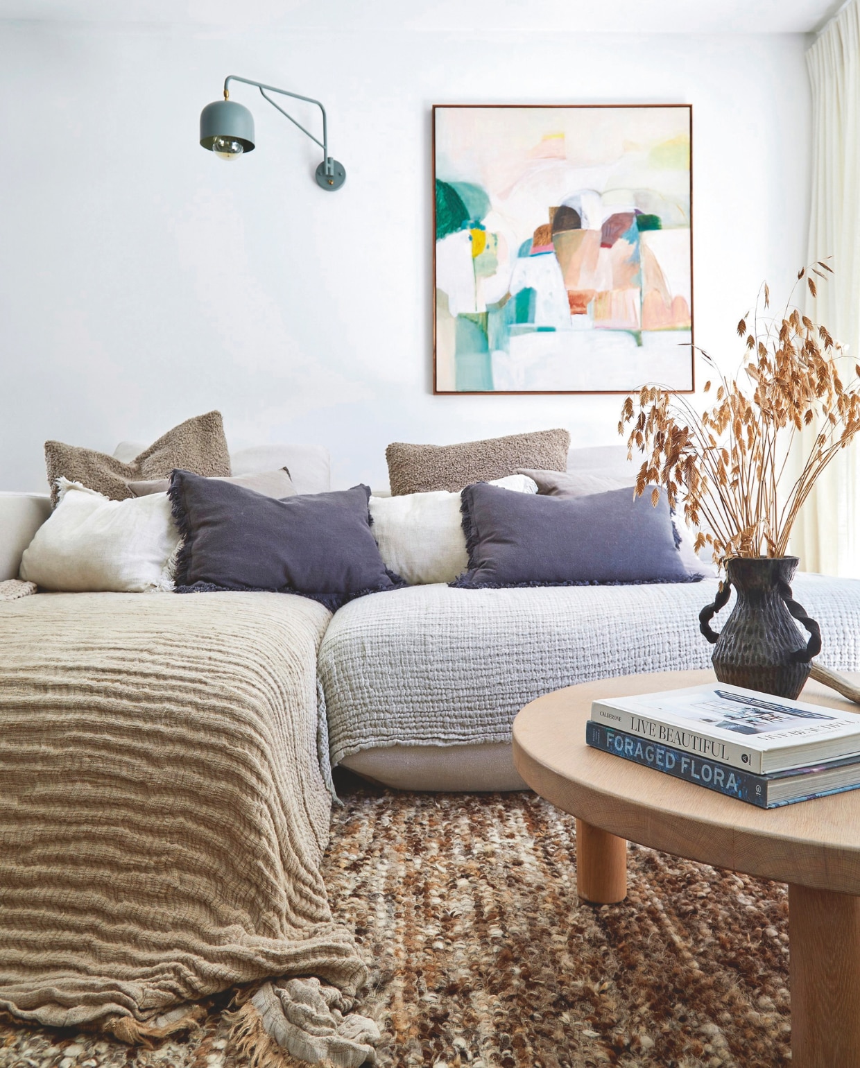 Bank La Grange Interiors. Muurlamp Acrobat van Acre Studio. Kunstwerk Laurinda Belcher. Vloerkleed, kussens en plaids Weylandts. Koffietafel Autumn and Oak. Vaas gemaakt door Janine voor Foraged. vtwonen 02-2026 | styling Sven Alberding | fotografie Greg Cox