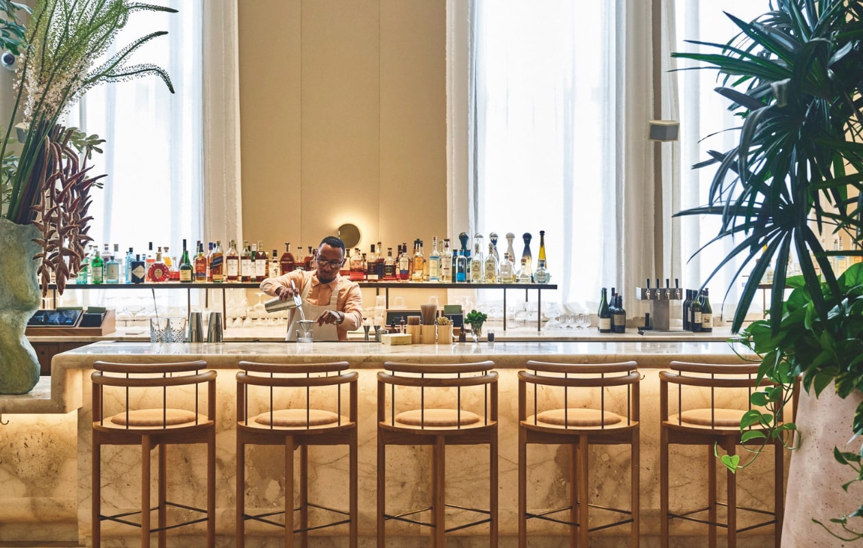 Plafondhoge ramen overspoelen de lobby met natuurlijk licht. Het interieur bevat onder meer marmeren bijzettafels van gerecyclede stenen uit een Spaanse steengroeve, vintage meubels uit Spanje en op maat gemaakte geweven tapijten uit Portugal. Wandkunst van Regina Jestrow. vtwonen Design 01-2026 | fotografie The Shelborne By Proper