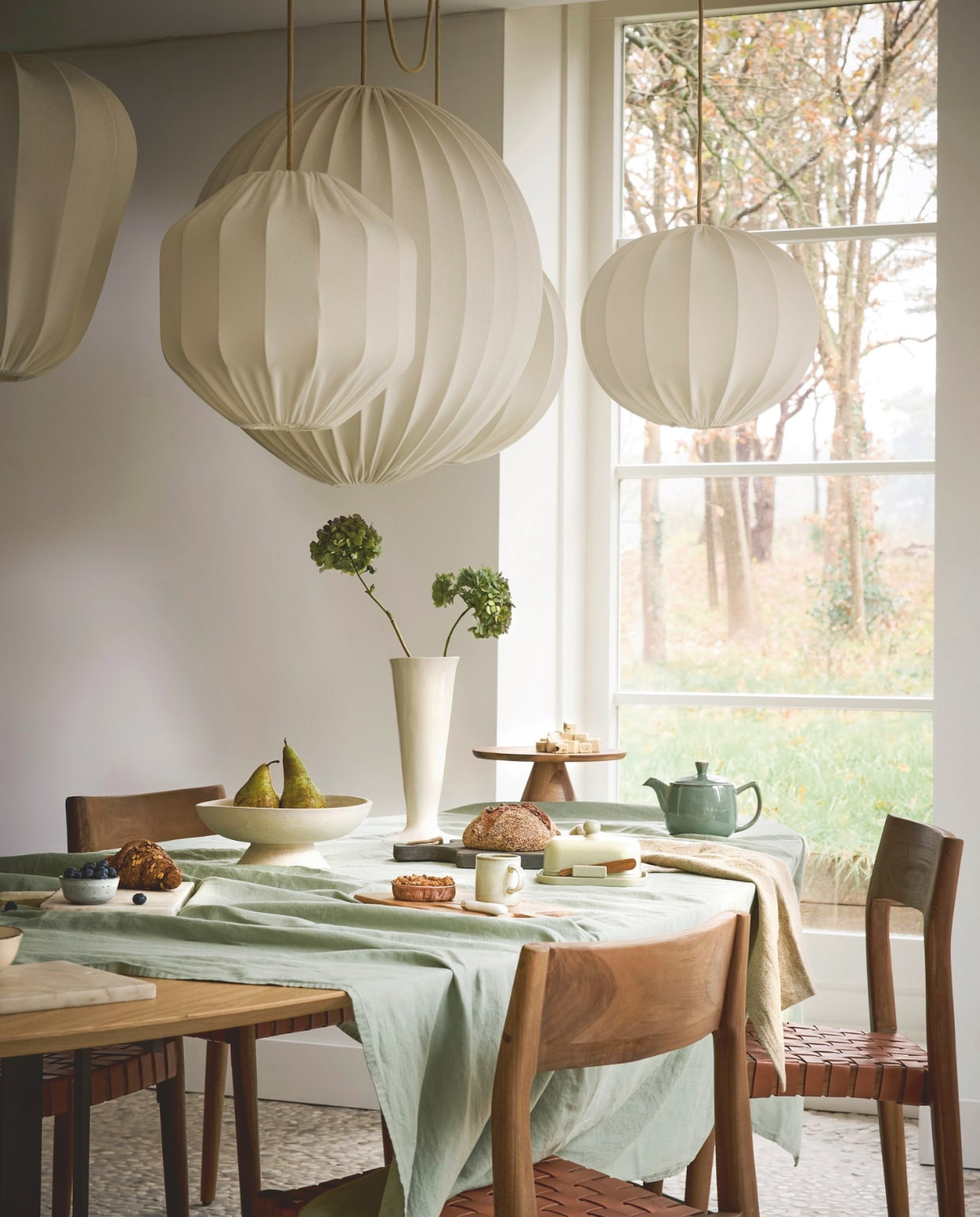 Lampverket hanglamp Myrull, draadframe en biologisch katoen, sand, van 30 cm (Ø) € 186,- tot 80 cm (Ø) € 490,-, hanglamp Droppe, draadframe en biologisch katoen, sand, 50 cm (Ø) € 174,- (lampverket.se). Raw Materials eetkamerstoel Freya, teakhout en leer, bruin, 46x49x80 cm (bxdxh) € 349,-, schaal Pano, chalk € 41,95, Urban Nature Culture vaas Mette, keramiek, wit € 67,99 (shop.vtwonen.nl). Bonsoirs tafellaken, katoen, groen, 170x250 cm (lxb) € 95,- (bonsoirs.com). Dille & Kamille taartplateau, acaciahout € 24,95, botermes, roestvrij staal en porselein € 2,95, theepot, steengoed, groen € 19,95, schaaltje, steengoed, grijs € 2,95 (dille-kamille.nl). Aura Peeperkorn snijplank, hout € 33,-, theedoek, linnen € 19,- (aurapeeperkorn.com). Zara Home botervloot met mes, aardewerk en hout, groen € 17,99, mok antieke rand, porselein, groen € 3,99, snijplank, hout, lichtbeige € 22,99 (zarahome.com). Overig privébezit. vtwonen Landelijk 02-2026 | styling Sanne Ketelaar | fotografie Alexander van Berge