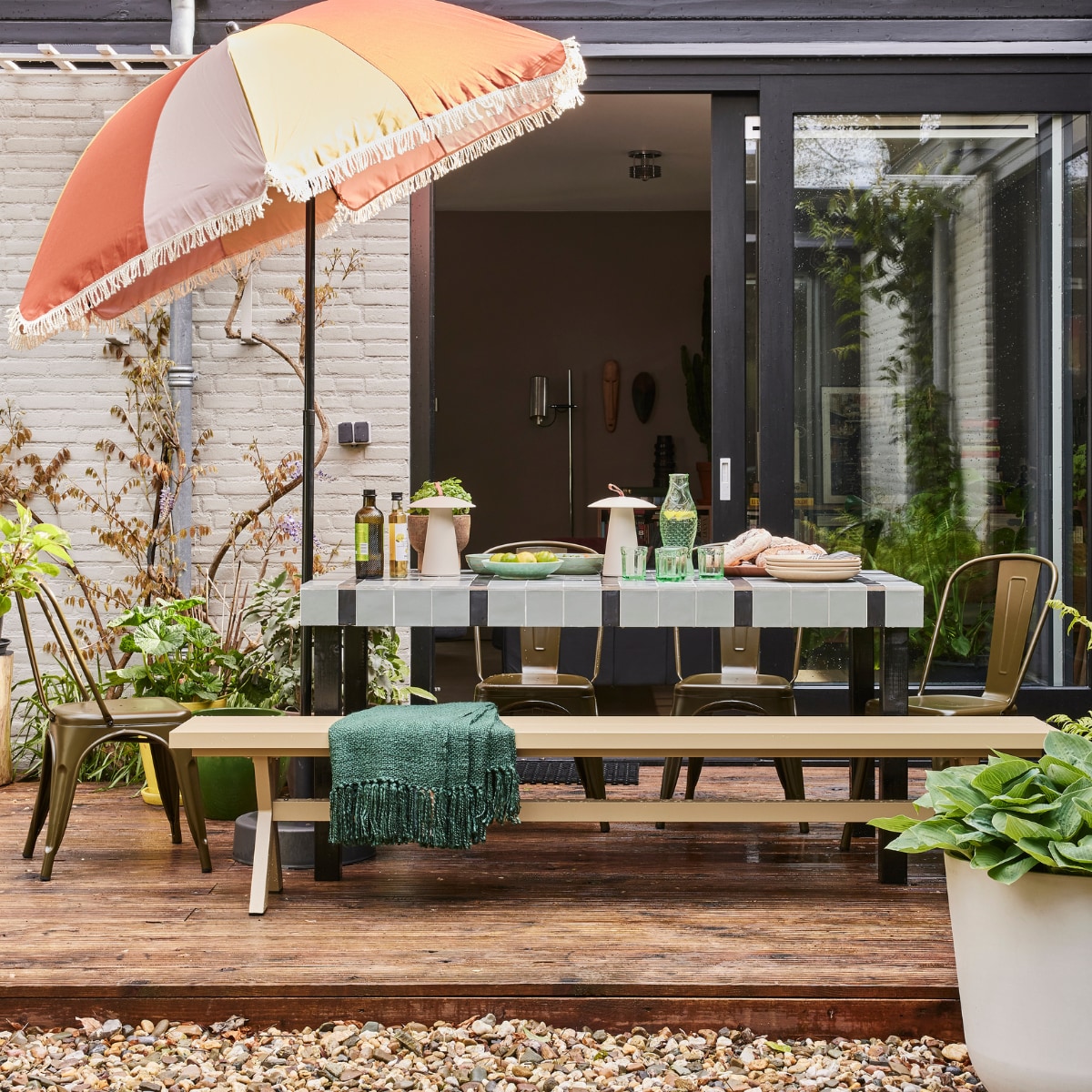 Tafel op maat gemaakt vanaf € 175,- (com-anders.nl), betegeld met vtwonen wandtegel Chop in Salvia, 10x10 cm € 79,90 per m² en wandtegel Chop Nero, 5x15 cm € 79,90 per m² (douglasjones.nl). Parasol € 49,99 (karwei.nl). Aluminium bank Delta van Exotan, 220x35x45 cm (lxbxh) € 299,-, plaid van Malagoon in Forest Green, 125x150 cm € 49,99, witgrijze plantenpot van Ecopots, 55 cm (Ø) € 99,- (vtwonen.nl/shop). Op tafel: draadloze lamp Ara van Nordlux € 129,95, viskaraf en glazen van Paola Navone voor Serax € 95,50, zeegroene saladeschaal, € 33,50 en dinerborden by fonq € 49,99 per 4 (shop. vtwonen.nl). Eetkamerstoel Afternoon van vtwonen € 79,95 (basiclabel.nl). Overig privébezit. vtwonen tuinspecial 01-2025 | styling Fietje Bruijn