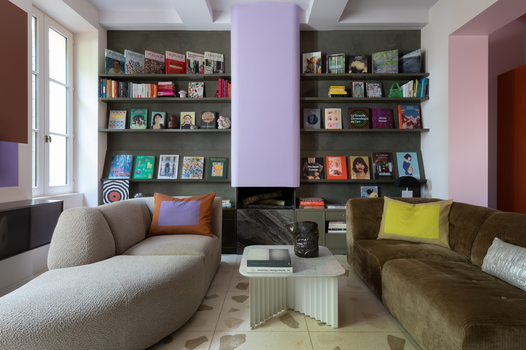 Centrale open haard in lavendelkleur geschilderd. Modulaire boekenkast ontworpen door Marine voor Montana Furniture. Modulaire banken van HKliving met kussens van Hay, HKliving en Ram Collection van Ana Kraš. Koffietafel Plec van Antoni Pallejà Office voor RS Barcelona met marmeren blad. Vaas-sculptuur van Victor. vtwonen Design 05-2026 | styling Francesca Sironi | fotografie Monica Spezia | productie Living Inside
