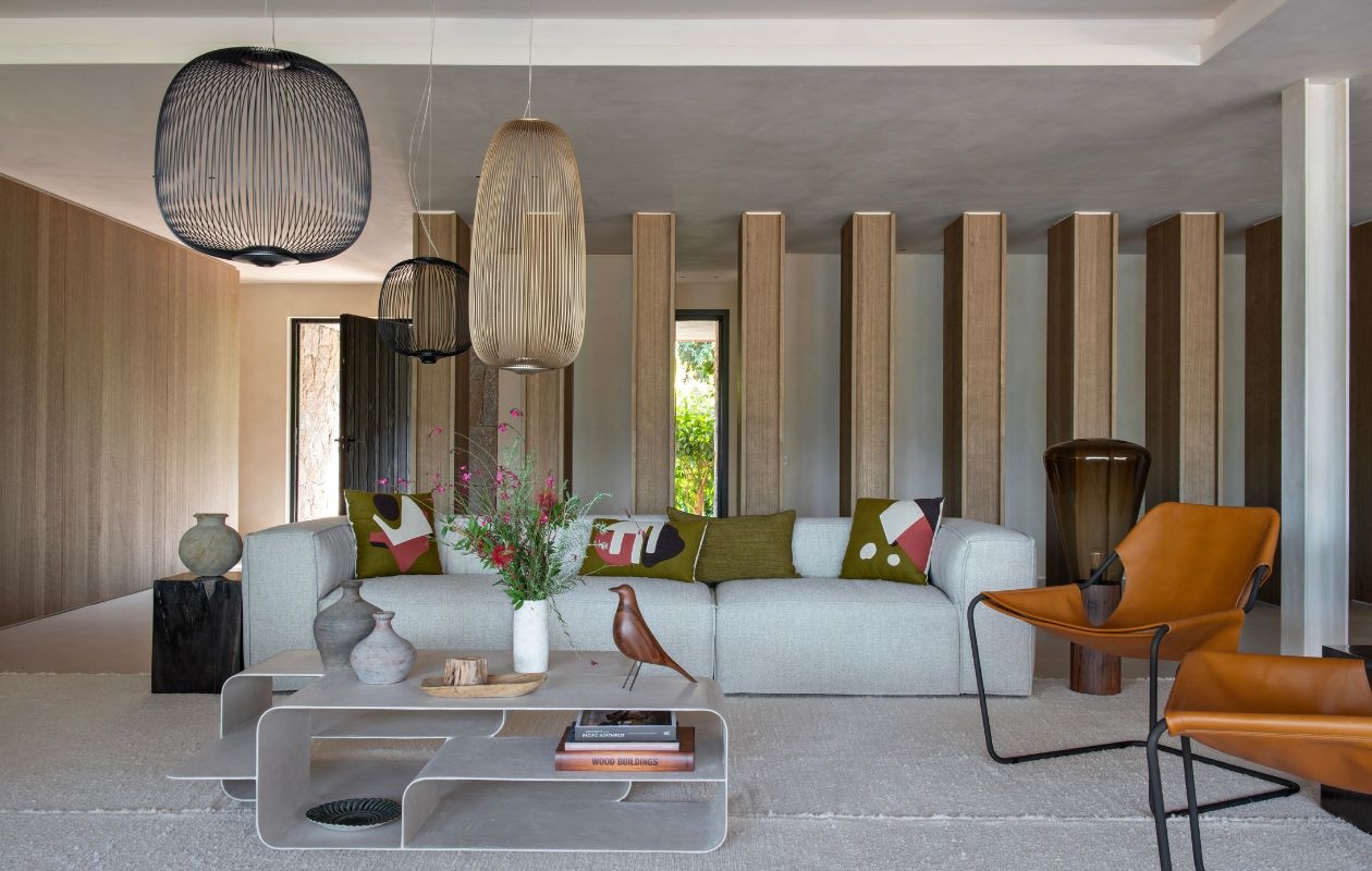 Woonkamer met zuilen van geborsteld en gebeitst eiken. Bank Slots Décoration. Kussens Maison de Vacances en Raphaëlle Malbec. Gerecycled katoenen tapijt Saudade Design. Hanglampen Foscarini. Bijzettafel van versteend hout. Vloerlamp Brokis. Salontafel L’Ocrier Studio. Notenhouten vogel Charles en Ray Eames. Vazen Ino Rive Droite. vtwonen Design 02-2026 | styling Laurence Dougier | fotografie Nicolas Mathéus | productie Living Inside