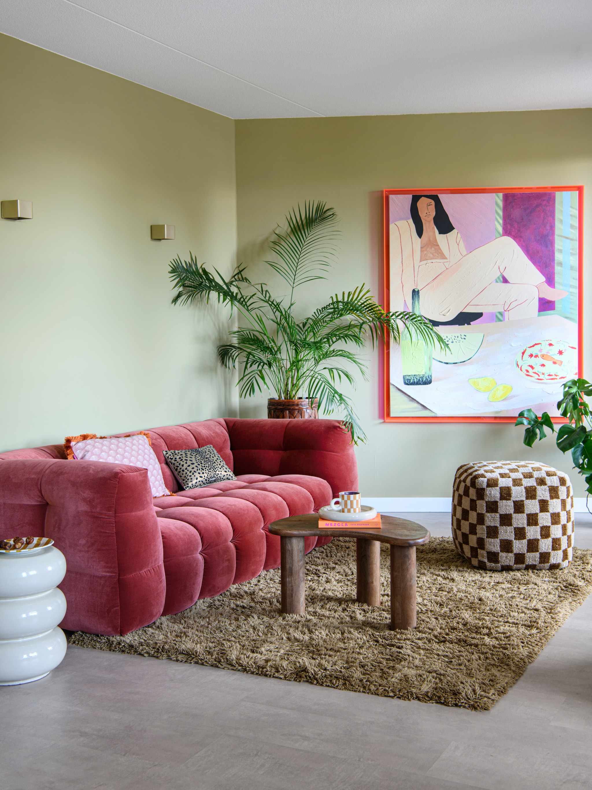 Vloer Karwei. Muurverf Puur Helmgras van Flexa. Bank Sofacompany. Poef, vloerkleed en koffiekop Loods5.Salontafel Home67. Bijzettafel en schotel kringloop. Kunstwerk HKliving. Wandlampen Luminize. Grote plant De Groene Brigade. Sierkussens Enkel Art Studio x Loods5 en HKliving.
