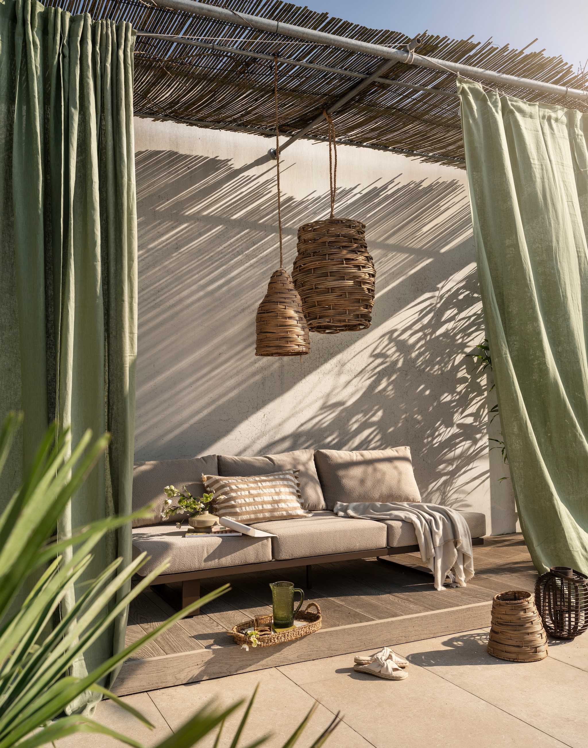 Woood tuinbank Bear 3-zits, outdoor stof en aluminium, bruin, 210x87x80 cm (bxdxh) € 999,- (shop.vtwonen.nl). Gommaire hanglamp Irving Xl, rotan, naturel € 295,-, hanglamp Nest, rotan, naturel € 199,-, mand Nest groot, wicker en pitriet, naturel € 60,- (gommaire.com). Ikea gordijnen Dytåg, katoen en linnen, grijsgroen, 300x145 cm (lxb), set van 2 € 69,99 (ikea.nl). Zara Home gestreepte kussenhoes, katoenmix, beige, 60x40 cm (lxb) € 39,99, deken met franje, katoenmix, ecru, 180x240 cm (lxb) € 39,99, glazen kan, reliëfmotief, glas, groen € 25,99 (zarahome.com). H&m Home kleine vaas, keramiek, beige € 14,99, ovaal dienblad, zeegras, naturel € 24,99, espadrilles met gedraaid detail, beige € 14,99 (hm.com). Kwantum lantaarn Bradford, bamboe, bruin € 19,99 (kwantum.nl). Overig privébezit. vtwonen Special 02-2026 | styling Sanne Ketelaar | fotografie Stan Koolen