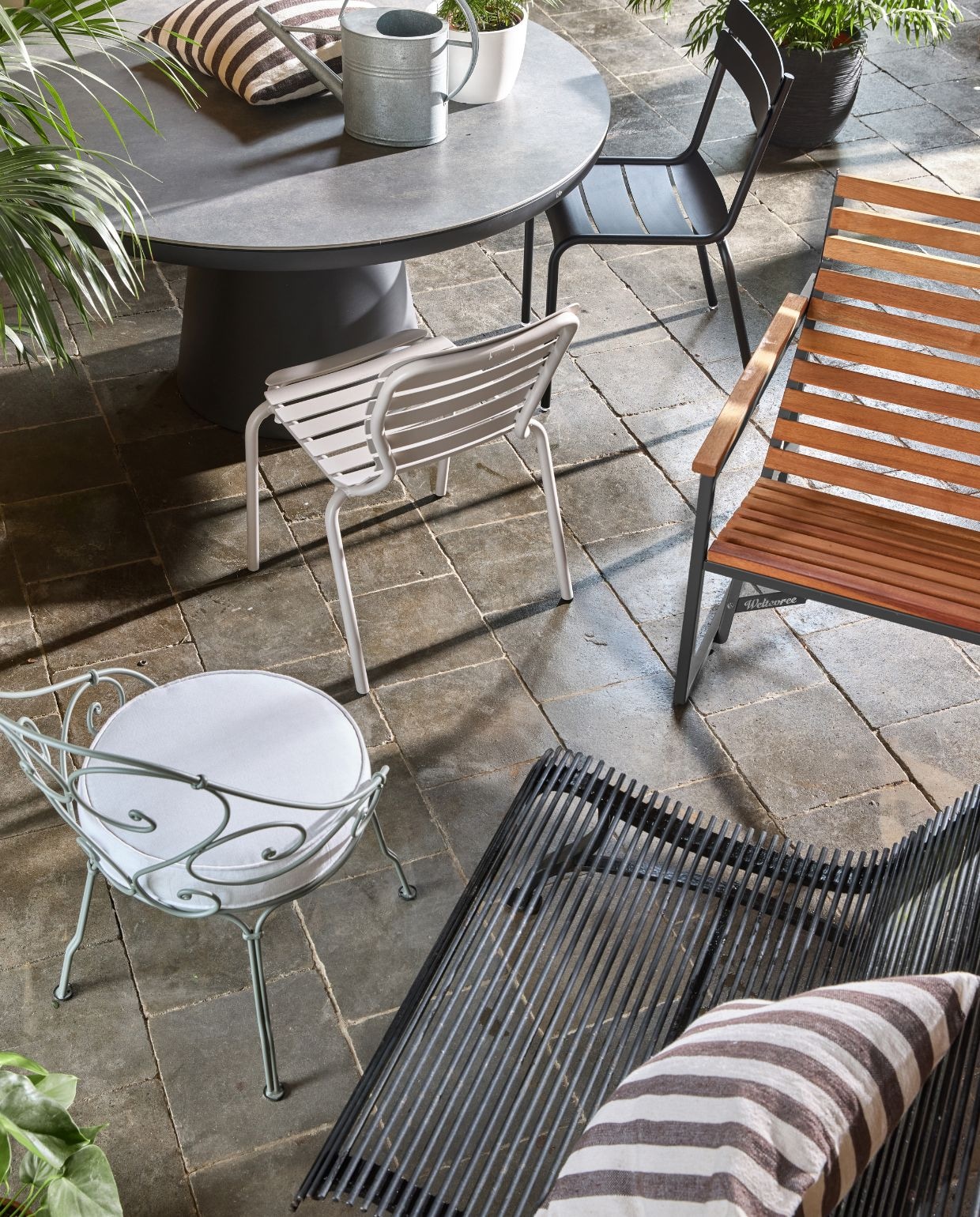 Life Outdoor Living eettafel Algarve, aluminium, lava, 150 cm (Ø) vanaf € 1099,- (lifeoutdoorliving.com).Weltevree bank Patiobench, metaal en hout, zwart € 995,- (weltevree.eu). Fermob draadstoel 1900 Cabriolet, met kussen, staalen stof, grey clay en off-white€ 525, stoel Luxembourg, aluminium, liquorice € 269,- (dacks.nl). Zuiver tuinstoel Vondel, aluminium, wit  € 269,- (zuiver.com). Household Hardware kussen, katoen, bruin/wit € 69,95 (householdhardware.nl). Intratuin gieter, zink € 24,99, Capi Europe pot met ribbels, zwart€ 139,-, Ecopots pot Oslo, wit € 25,95 (intratuin.nl). vtwonen TUIN 2026 | styling Marlies Does | fotografie Peggy Janssen