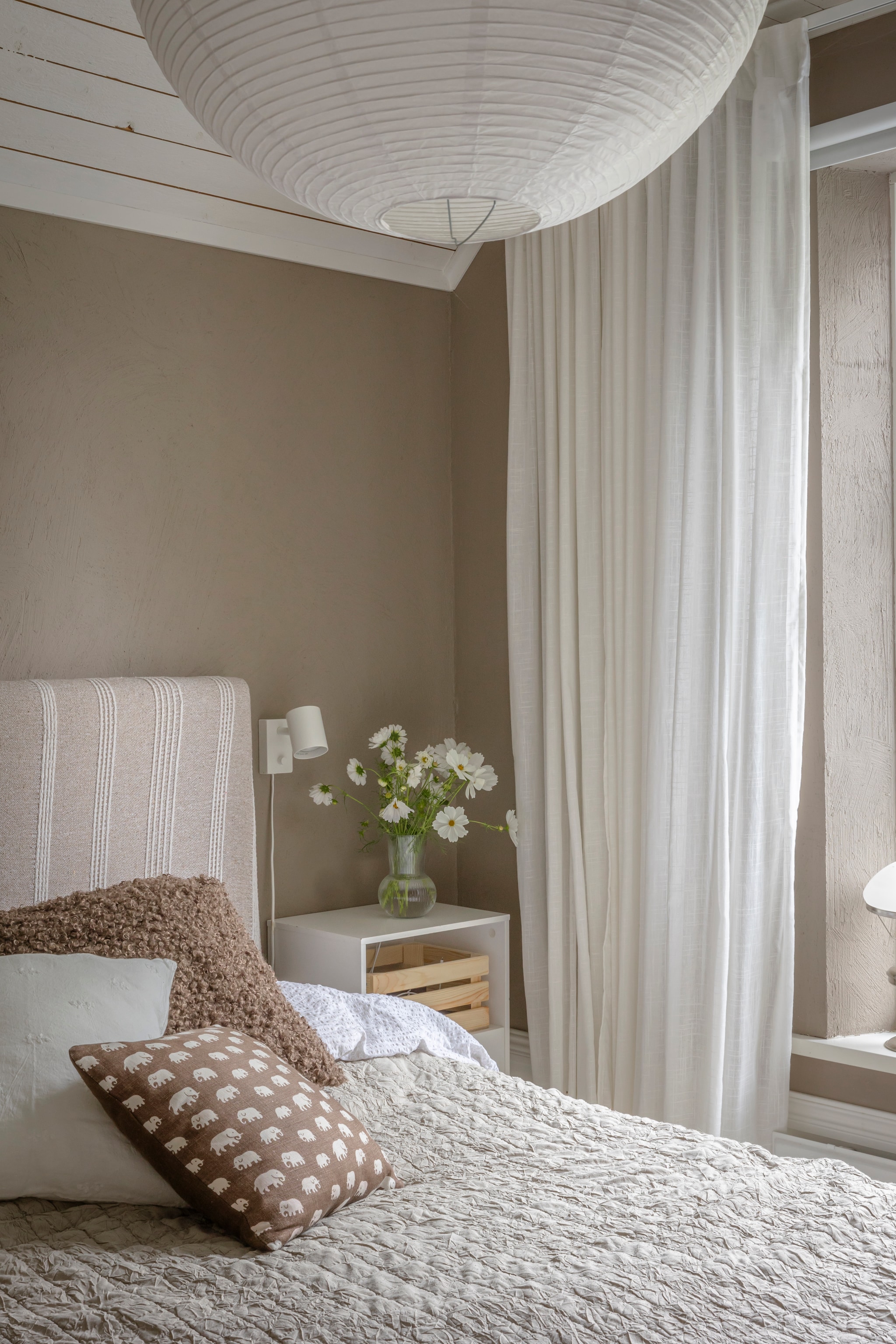 Muurverf Muskatnøtt van Jotun. Gordijnen, hanglamp, vaas, wandlamp en krat Ikea. Beddengoed Rusta. Hoofdbord zelf gemaakt en bekleed met overgebleven stof van Rusta. Kussens Svenskt Tenn en Lilla Stenhuset. Nachtkastje Ellos. vtwonen Landelijk 06-2026 | styling Anna Bjelkholm | fotografie Anna Karlsson