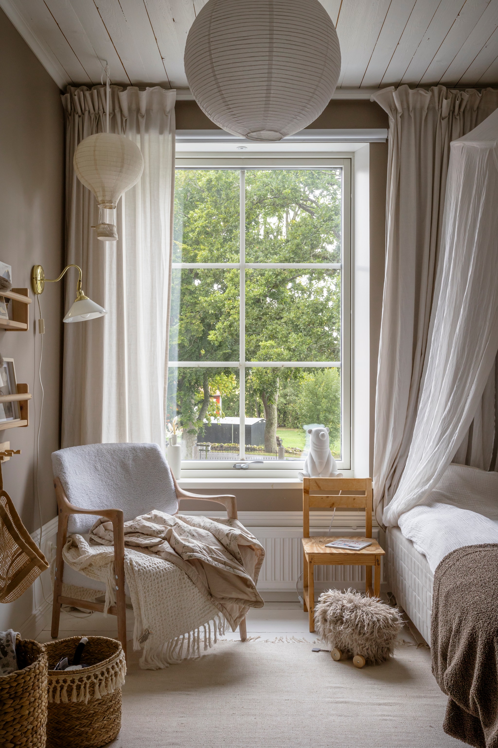 Muurverf Muskatnøtt van Jotun. Oorspronkelijke vloer, wit geschilderd. Bed, vloerkleed, gordijnen,  hanglamp, wandplanken en gehaakte plaid Ikea. Gebloemde plaid Garbo & Friends. Beddengoed Lilla Stenhuset. Klamboe en speelgoedschaap Jollyroom. Luchtballon-lamp, wandlamp en kleine stoel kringloop. Manden Hemtex. Fauteuil veiling.  vtwonen Landelijk 06-2026 | styling Anna Bjelkholm | fotografie Anna Karlsson