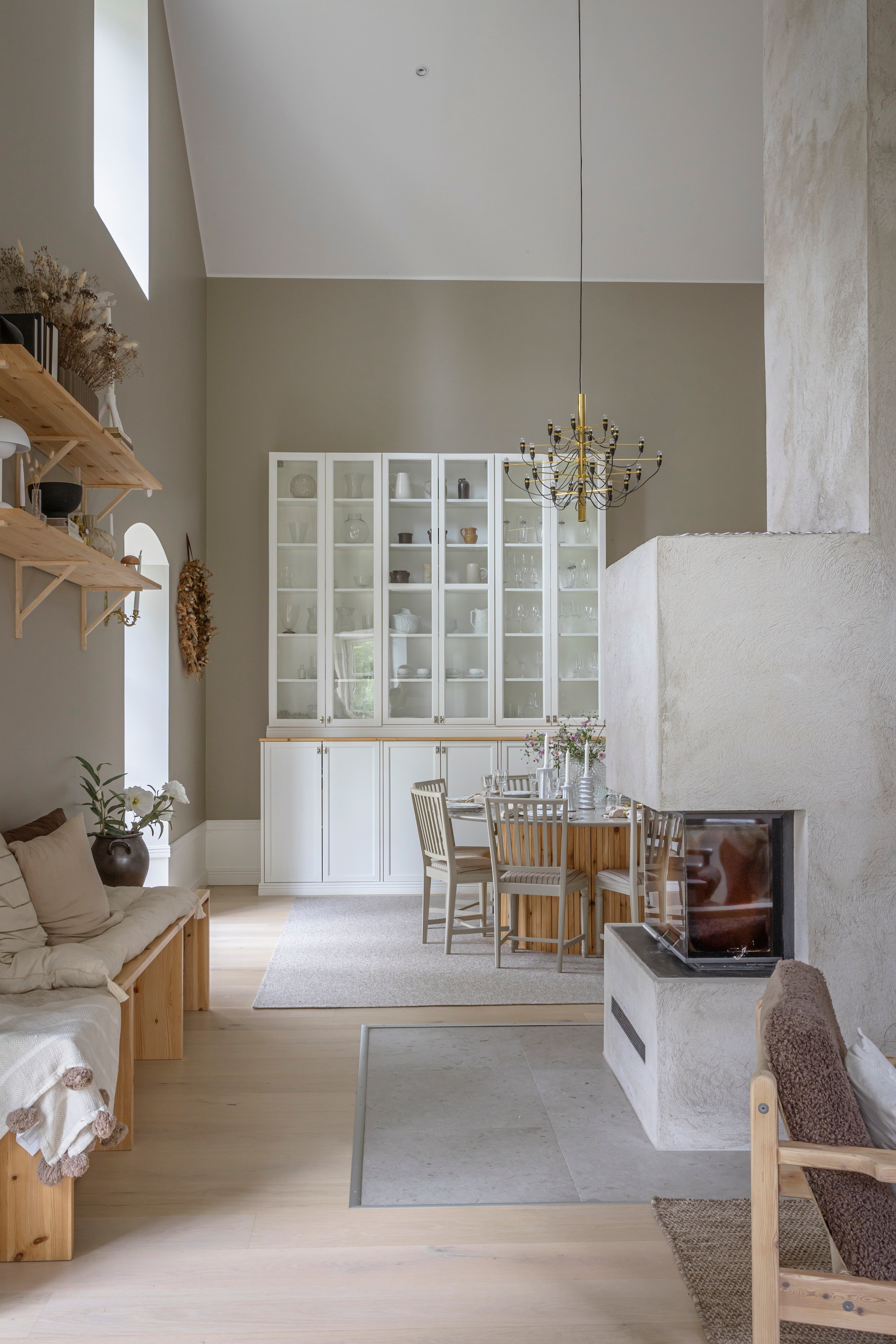 Muurverf s3005-y20r van Jotun. Open haard Contura, metselwerk eromheen zelf gedaan. Vloer gewit eiken van Bjelin. Vloertegels kalksteen. Houten bank, tafel en wandkast zelf gemaakt. Zitkussens H&m. Sier-kussens Lilla Stenhuset en Ernst Form. Vloerkleed Stuvbutiken. Stoelen en servies veiling. Kroonluchter Flos.Wandplanken Biltema. vtwonen Landelijk 06-2026 | styling Anna Bjelkholm | fotografie Anna Karlsson