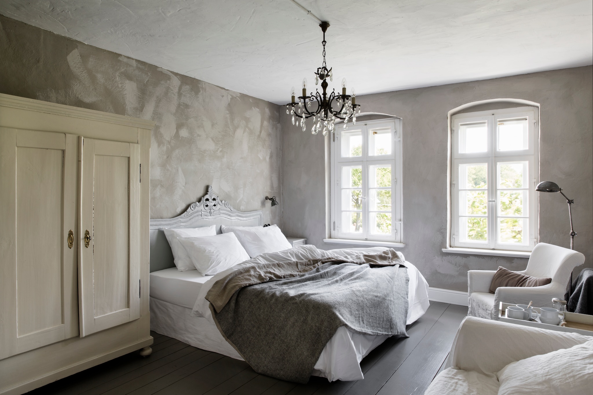 Hoofdbord Old Basics. Bed en beddengoed Fbf bed&more. Linnenkast en kroonluchter antiek. Fauteuils en vloer-lamp Loberon. Op de muur kalkverf, zelf gemengd met ecologisch pigment, beide Kreidezeit. Vloerverf Disbon 404 van Caparol, gemengd in de oorspronkelijke kleur van de vloer. vtwonen Landelijk 06-2026 | styling Miriam Hannemann | fotografie André Reuter