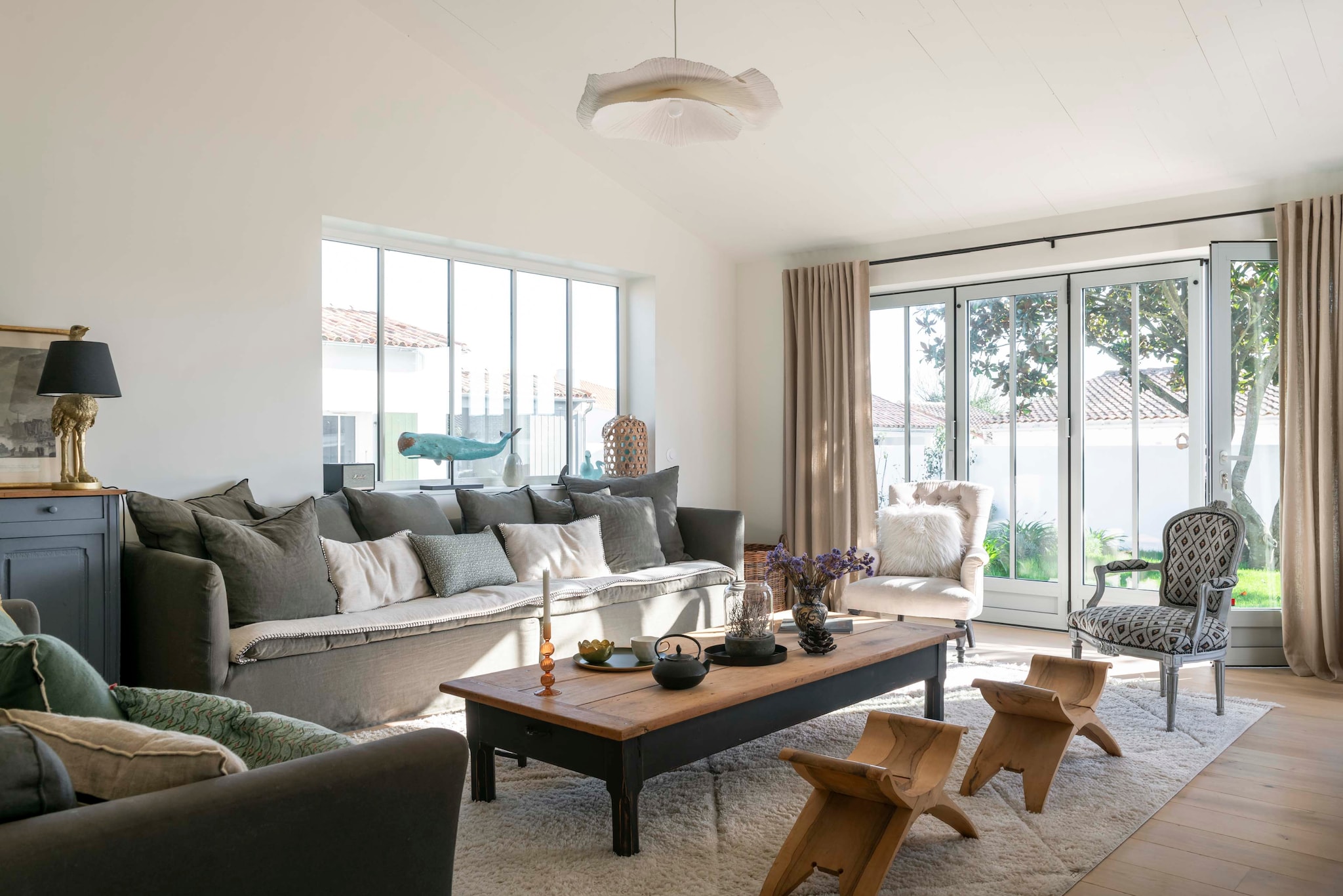 Bank Maison de Vacances. Fauteuils uit de familie. Saramaka-krukken uit Frans-Guyana meegebracht. Salontafel en dressoir Brocante Marchand d’Oublis. Kussens La Redoute. Hanglamp Zara Home. Vloerkleed berber uit Marokko. Tafellamp Kare Design. Walvissculptuur lokale boetiek Migration. Gordijnen Mondial Tissus. vtwonen Landelijk 06-2026 | styling Amandine Berthon | fotografie Julien Fernandez | productie Amandine & Jules