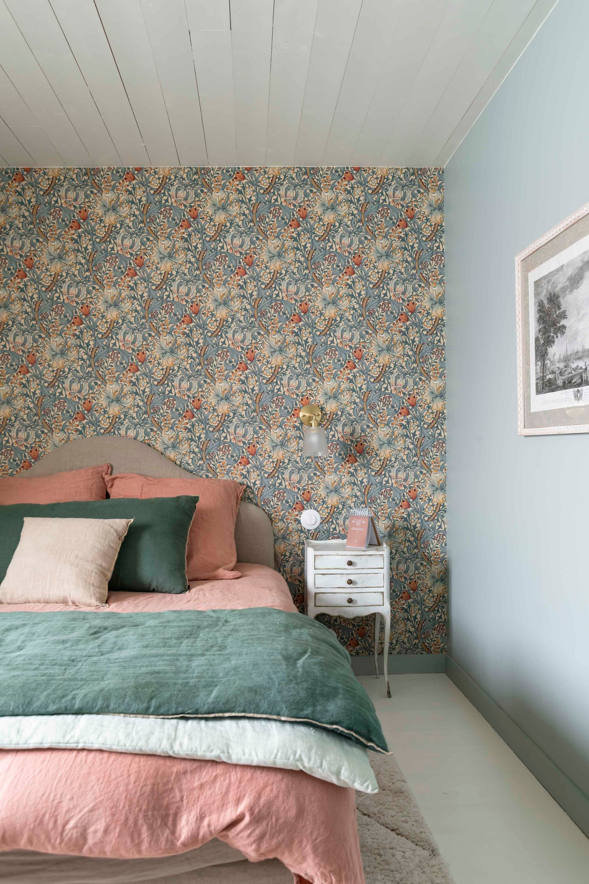 Behang Golden Lily van Morris & Co. Wandlamp Cyrillus. Bed vintage met opnieuw bekleed hoofdbord. Nachtkastje familiestuk. Beddengoed, gordijnen en kussens Haomy Textile. Gravure erfstuk. Berber vloerkleed uit Marokko. Wandkleur Light Blue van Farrow & Ball. vtwonen Landelijk 06-2026 | styling Amandine Berthon | fotografie Julien Fernandez | productie Amandine & Jules
