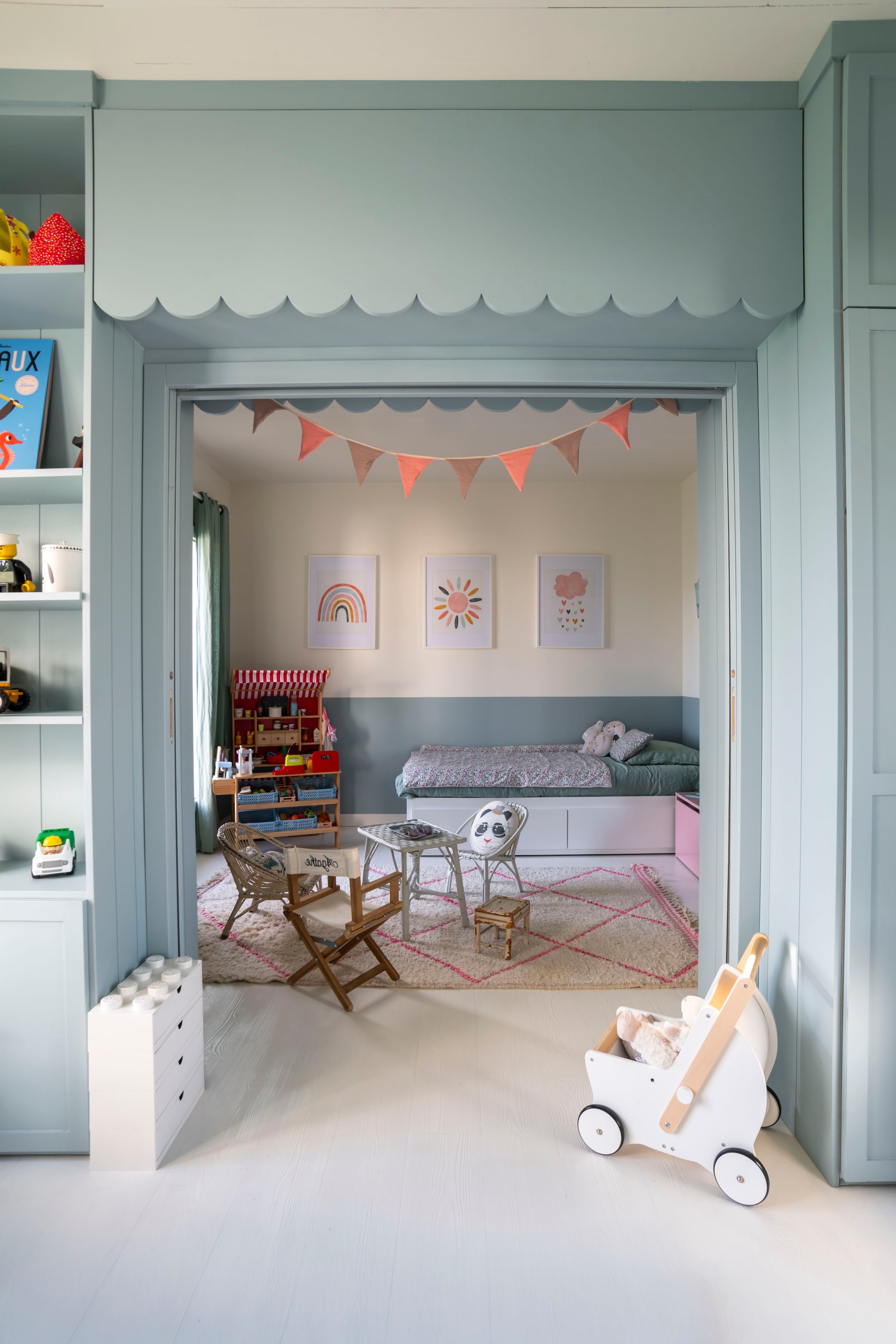 Schuifdeur omlijst door opbergruimte gebouwd door Pia Île de Ré. Berber vloerkleed uit Marokko. Houten speelgoed en winkeltje Howa. Kindermeubels vintage vondsten. Gordijnen Haomy. Bed Ikea. Beddengoed Linvosges. Illustraties Etsy. vtwonen Landelijk 06-2026 | styling Amandine Berthon | fotografie Julien Fernandez | productie Amandine & Jules