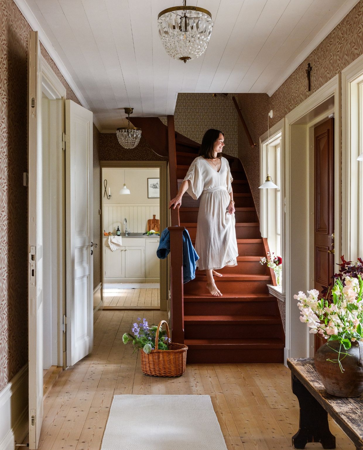 Nieuwe trapleuning in rustieke stijl, geschilderd met lijnolieverf Bränd Terra van Engwall & Claesson. Tapijt H&m Home. Bank Dakabom. Mand en vazen tweedehands. Bloemen uit eigen tuin: leeuwenbek, ridderspoor, Ammi majus en siergras. Behang Willow Russet van William Morris. Deuren en houtwerkgeschilderd met lijnolieverf Nougat 10% van Ottosson. vtwonen Landelijk 03-2026 | fotografie Rebecca Martyn