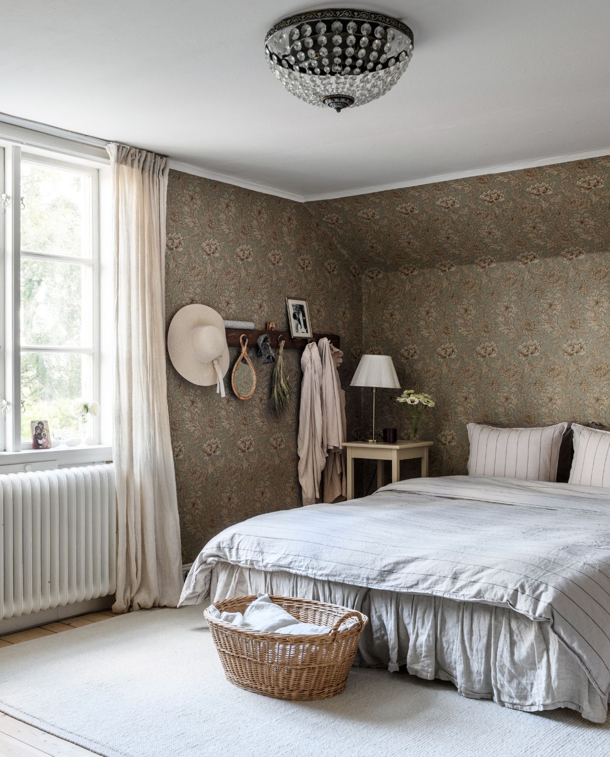 Dekbedovertrek Gripsholm Zweden. Linnen bedrok Ellos. Linnen gordijnen H&m Home. Mand tweedehands. Tapijt Himla. Behang Chrysanthemum van William Morris. Bloemen uit eigen tuin: zinnia’s. De kapstok hing er al, zelf geschilderd. Nachtkastje tweedehands, geschilderd met lijnolieverf. vtwonen Landelijk 03-2026 | fotografie Rebecca Martyn