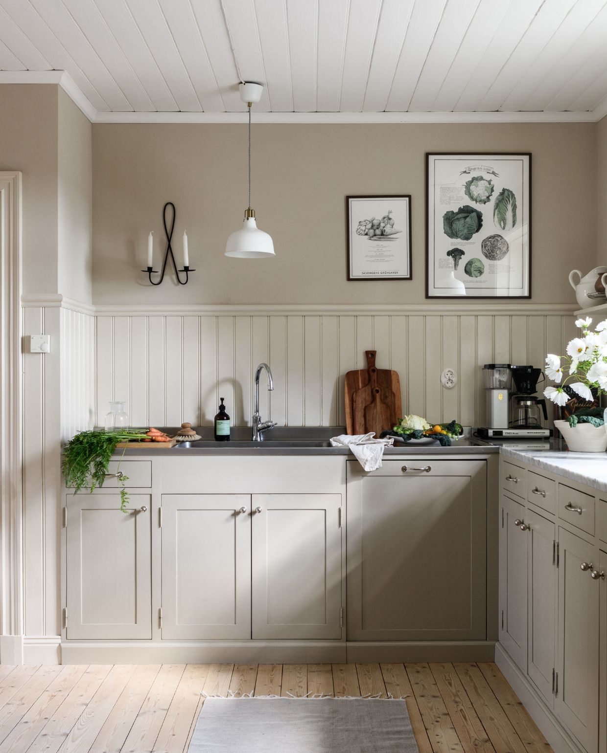 Keuken: Basis Ikea met deuren en sierlijsten van lokale timmerman, geschilderd met lijnolieverfSchweizer Grijs 25% van Engwall & Claesson. Lambrisering en lijstwerk, geschilderd met lijnolieverf Nougat 10% van Engwall & Claesson. Plank zelfgemaakt door Johannes. FornuisLofra. Lampen Ikea. Bloemen en groenten uit eigen tuin. Vazen en karaffen tweedehands. Tapijt Himla. Smeedijzeren kandelaar Boozt. vtwonen Landelijk 03-2026 | fotografie Rebecca Martyn