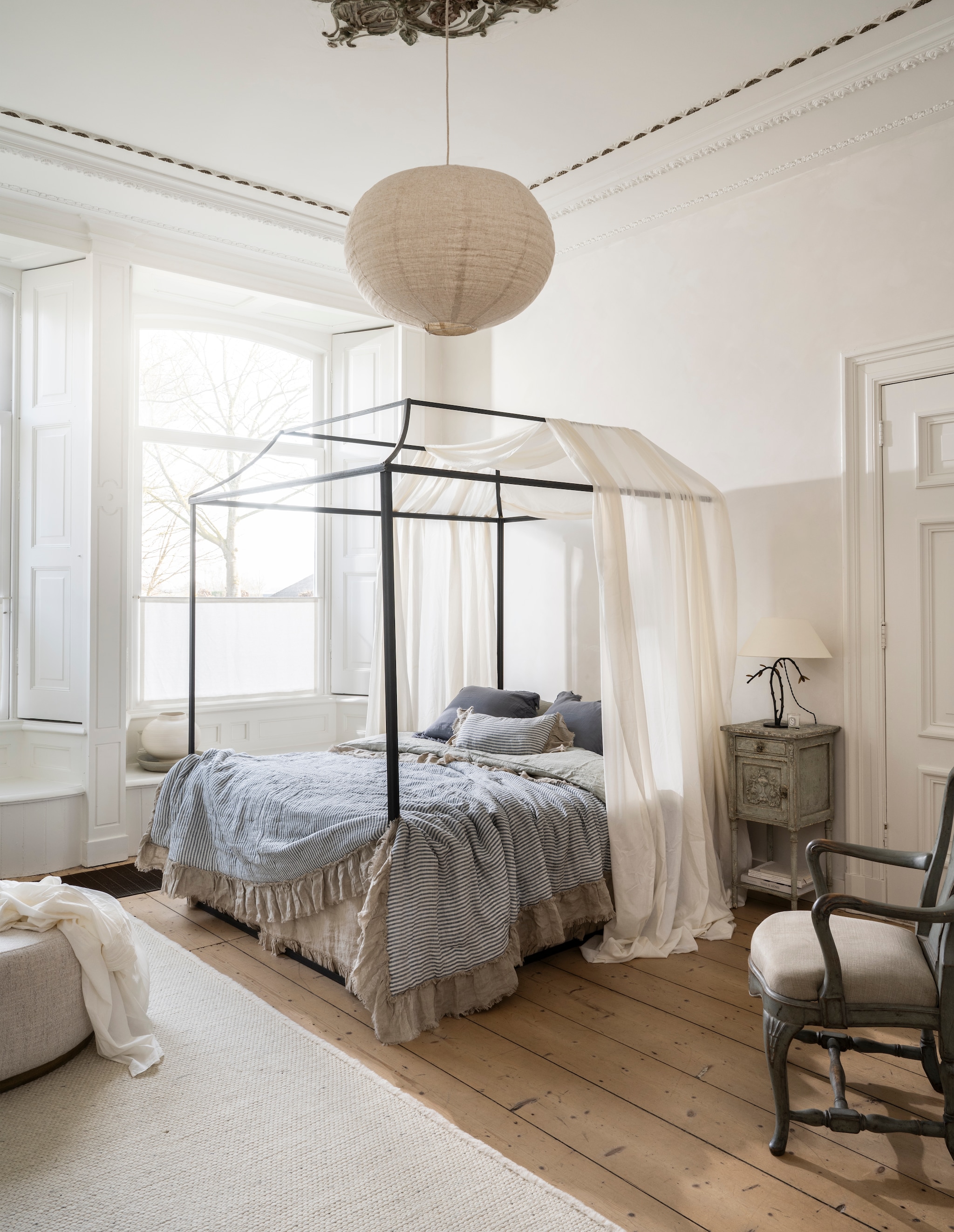 Zara Home hanglamp, linnen, 65x46 cm (Øxh) € 59,99, tafellamp Colin King, messing, zwart en wit, 36x17x29 cm (lxbxh) € 419,-, ochtendjas, katoen, ecru € 69,99 (zarahome.com). Piet Jonker Antiek Frans nachtkastje, hout, grijs patina, 38,5x38x83 cm (bxdxh), prijs op aanvraag, 19e-eeuwse Zweedse fauteuil, hout en linnen, 60x50x98 cm (lxbxh), prijs op aanvraag (pietjonker.nl). Rivièra Maison vloerkleed Crème de la crème, pebble beach, 290x200 cm (lxb) € 1265,40, poef Babylon, tweed, antique white, 91x43 cm (Øxh) € 599,- (rivieramaison.com). Urban Nature Culture vaas Camila, keramiek, wit, 31x27 cm (Øxh) € 89,99 (shop.vtwonen.nl). Combitex hemelbed, metaal, zwart, 200x160 cm (lxb), prijs op aanvraag (com-anders.nl). Loberon Quilt met ruches Alexane, linnen en katoen, blauw en wit, 260x240 cm (lxb) € 228,-, kussenhoezen met ruches Alexane, linnen en katoen, blauw en wit, set van 2 € 39,95 (loberon.nl). Yumeko dekbedovertrek en kussenhoezen, gewassen linnen, misty green € 359,- (shop.vtwonen.nl). Casa Comodo laken met ruches, linnen, classic sand € 224,95 (casacomodo.nl). vtwonen Landelijk 06-2026 | styling Kim van Rossenberg | fotografie Stan Koolen