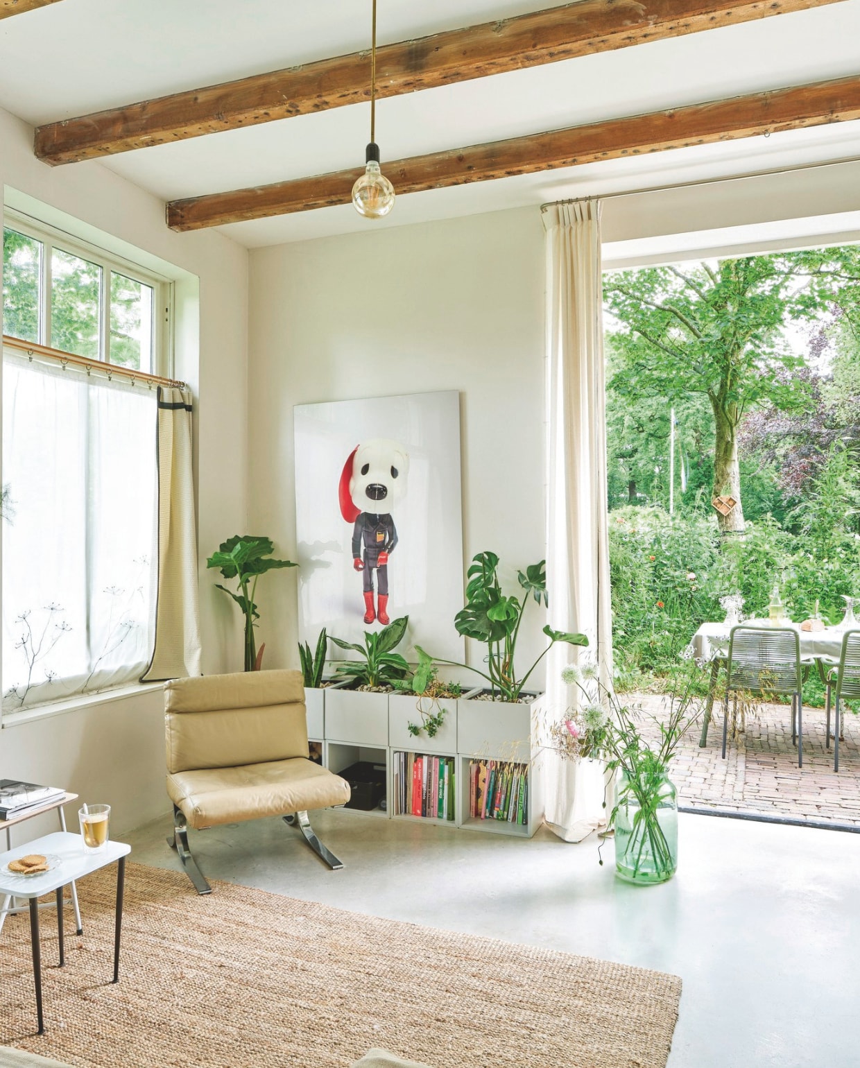 Vloer gevlinderd beton met vloerverwarming. Bijzettafels kringloop en Van Dijk & Ko. Vloerkleed en wandmeubel Ikea. Wandvaas Kastanje van Anneke Bruin. Gordijnen van kaasdoek met geborduurde schermbloemen, overgordijnen, linnen gordijnen bij openslaande deuren en lamp Brigit Vermeulen. Fauteuil Zo Goed als Oud. Kunstwerk The Indispensable van Ilse Vermeulen. Glazen vaas Van Dijk & Ko. Tuinstoelen Karwei. Tuintafel Tavira van Gamma. vtwonen 03-2025 | fotografie René van der Hulst