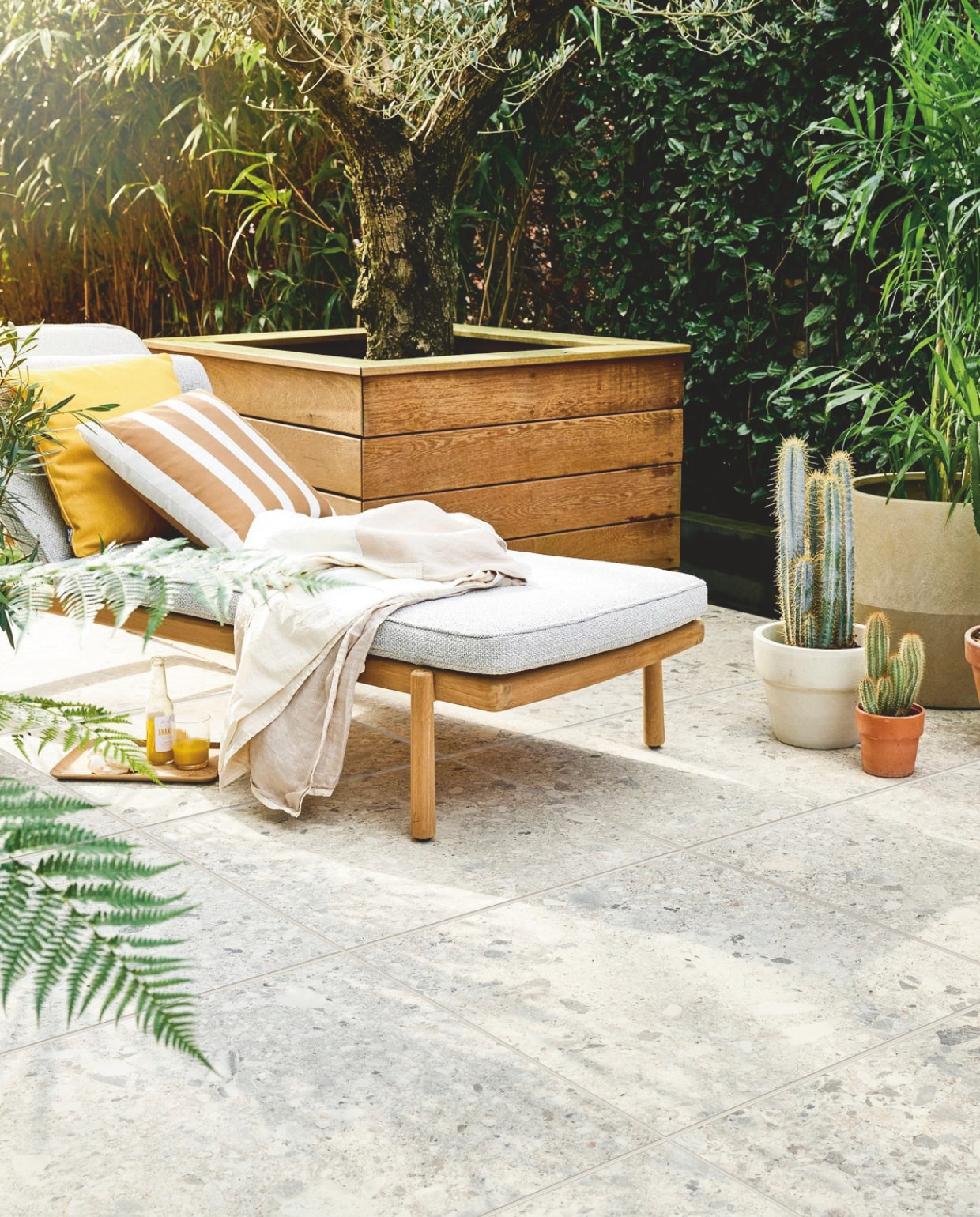 Tuintegel Solostone Uni Composite Mix Beige, 70x70 cm, per m² € 74,50, ook verkrijgbaar in Mix Grey. M.m.v. dacks.nl (ligbed met matraskussen Pure Sofa van Tribù), intratuin.nl (geel kussen, gestreept kussen, terracotta bloempot), vtwonen.nl/shop (plaid, witte bloempot van Serax), zarahome.com (dienblad, glas). Overig privébezit. vtwonen 05-2024 | styling Marlies Does | fotografie Sjoerd Eickmans