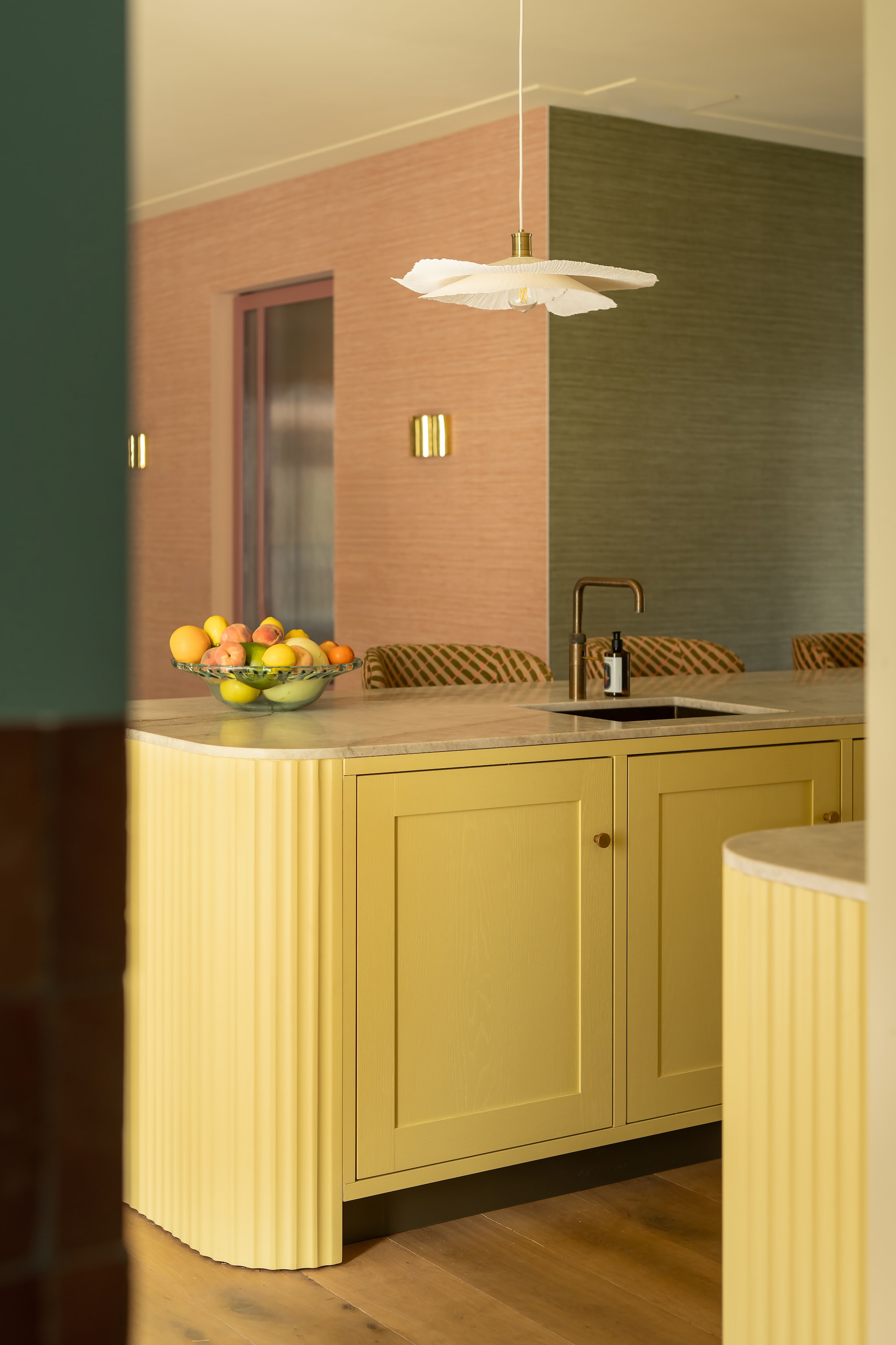 Keuken maatwerk met aanrechtblad van Taj Mahal-kwartsiet. Barstoelen Kave Home, bekleed met stof Stripes 10966.56 van Nobilis. Hanglampen Zara Home. Kleur keukenblokken Gervaise Lemon, plafond Light Stone, wand Hound Lemon van Farrow & Ball. Behang Arte Essentials Tangram Caprice Linen 24004. Wandlamp Sézane. vtwonen Design 06-2026 | styling Juliette de Dreu | fotografie Lotte van Uittert