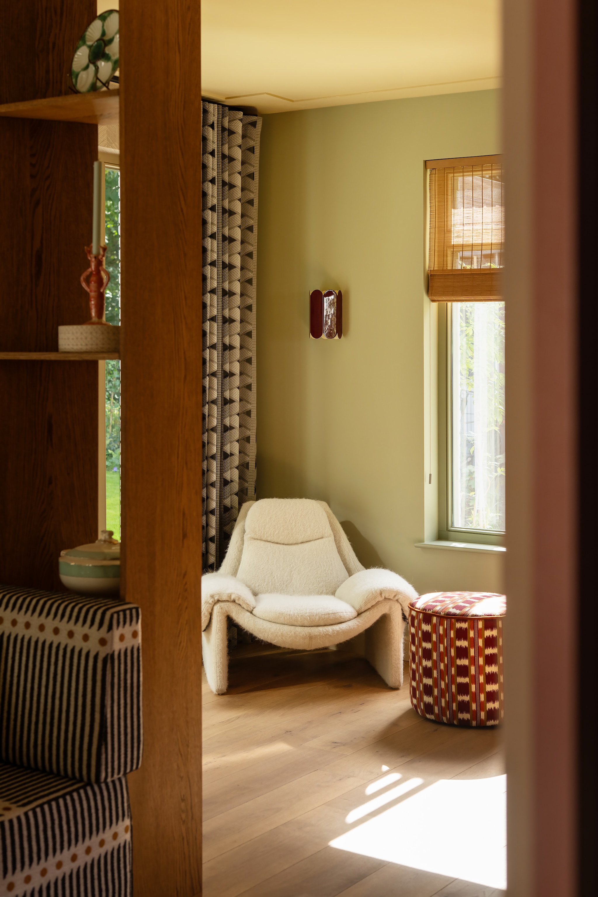Gordijnen Toppoint. Wandlamp Hay. Fauteuil P60 Saporiti via Nu-Design Meubelatelier, bekleed met stof Yeti van Pierre Frey. Poef Studio Habib, bekleed met stof van Manuel Canovas. Verfkleur wand Vert de Terre, raamkozijnen Lichen, kozijnen pui Light Stone van Farrow & Ball. Roomdivider en eetkamerbank gemaakt door Marijn Interieurs, bekleed met stof van Nobilis. Vloer van verouderde eikenhouten planken via eikenvloerdelen.nl. vtwonen Design 06-2026 | styling Juliette de Dreu | fotografie Lotte van Uittert