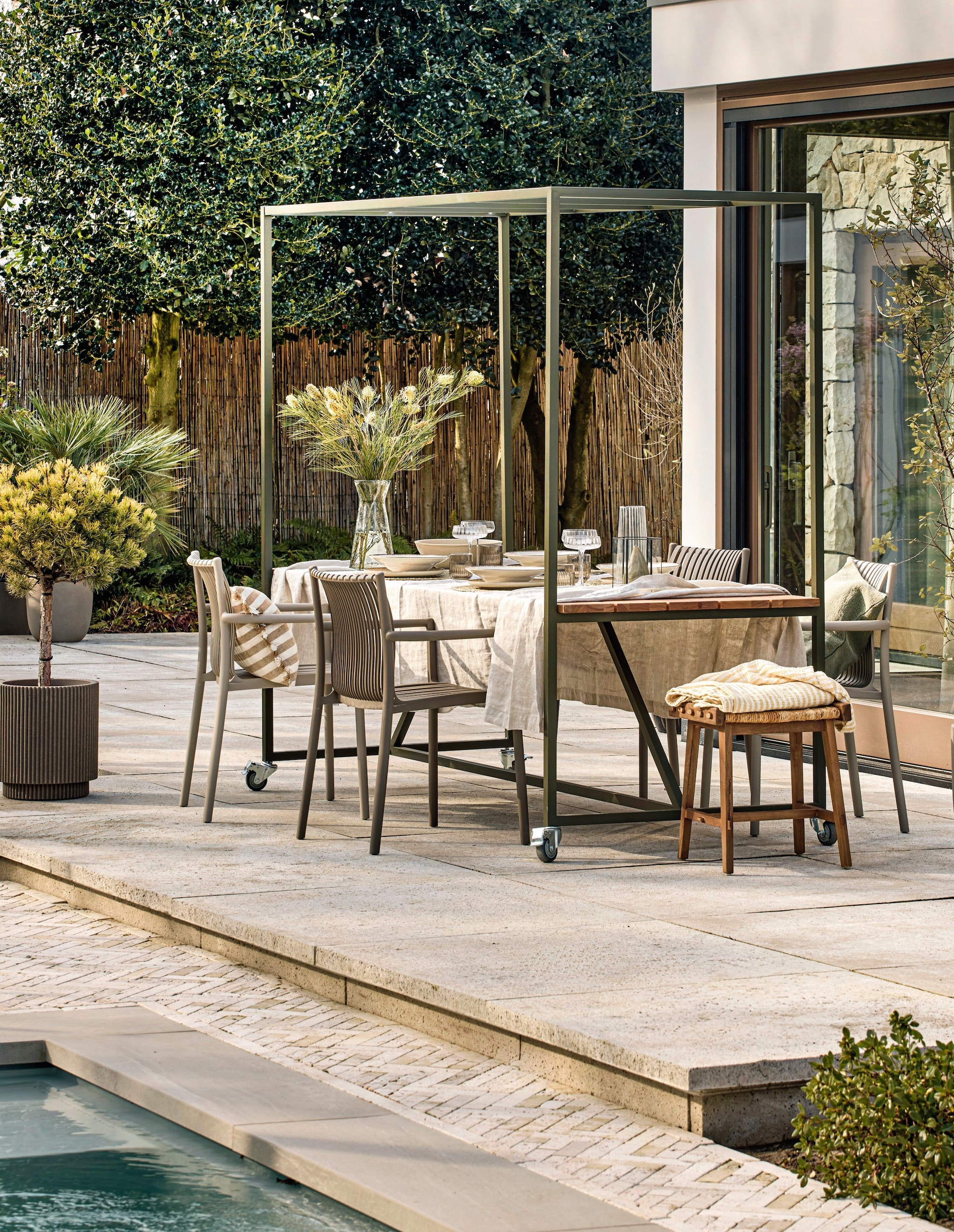 vtwonen eettafel Blue Sky op wielen, metaal en hout, dust, 220x97x200 cm (lxbxh) € 599,-, vtwonen tuinstoel Easy Going, kunststof, zand en bruin, 56x57x84 cm (bxdxh) € 99,95.M.m.v. zarahome.com (kussens, plaid, vaas, servies), loods5.nl (glazen kan, cocktailglazen, bruine glazen), hetkabinet.nl (kruk).