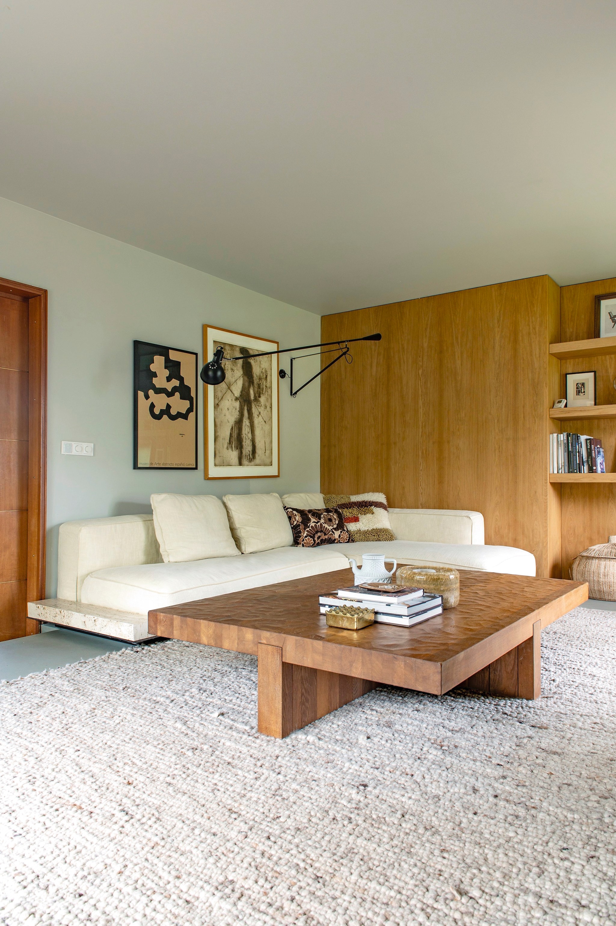 Salontafel van Linteloo via Whoppah. Bank Grandemare van Flexform. Wandlamp 265 van Floss. Kunstwerk rechts Werner Moonen. Kunstwerk links lithografie op kraftpapier Sin Titulo van Eduardo Chillida. Vloerkleed, wollen kussen en windlicht Ony via Bynord. Bloemkussen HKliving. Theepot Sargadelos. Kleur wand en plafond Old Flax van Pure & Original.