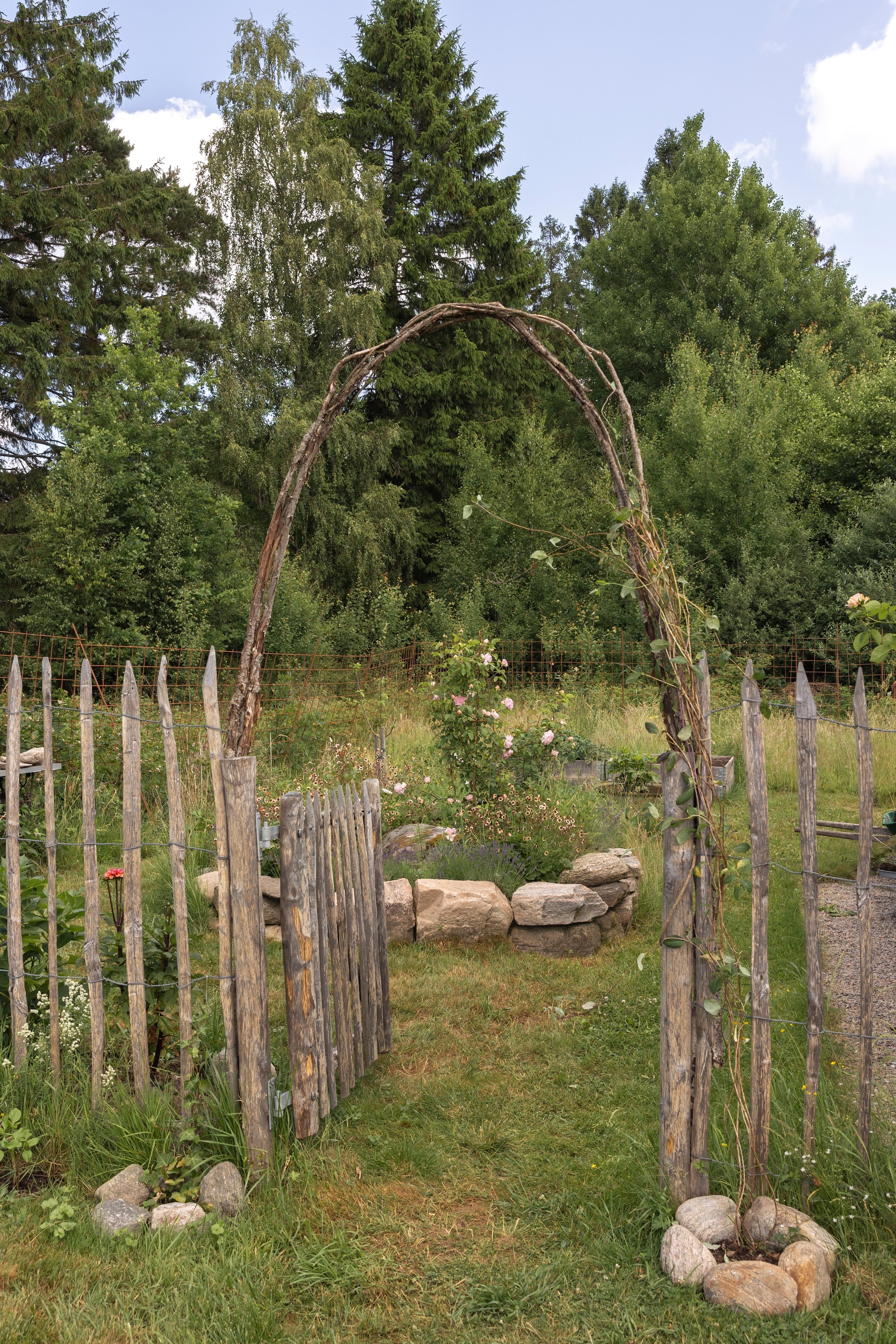 De moestuin is afgezet met een kastanjehouten omheining. Soortgelijke omheining Adéquat. Om de boog bloeit straks kamperfoelie. vtwonen Landelijk 07-2026 | productie Sarah MacAfie | styling Anna Bjelkholm | fotografie Anna Karlsson
